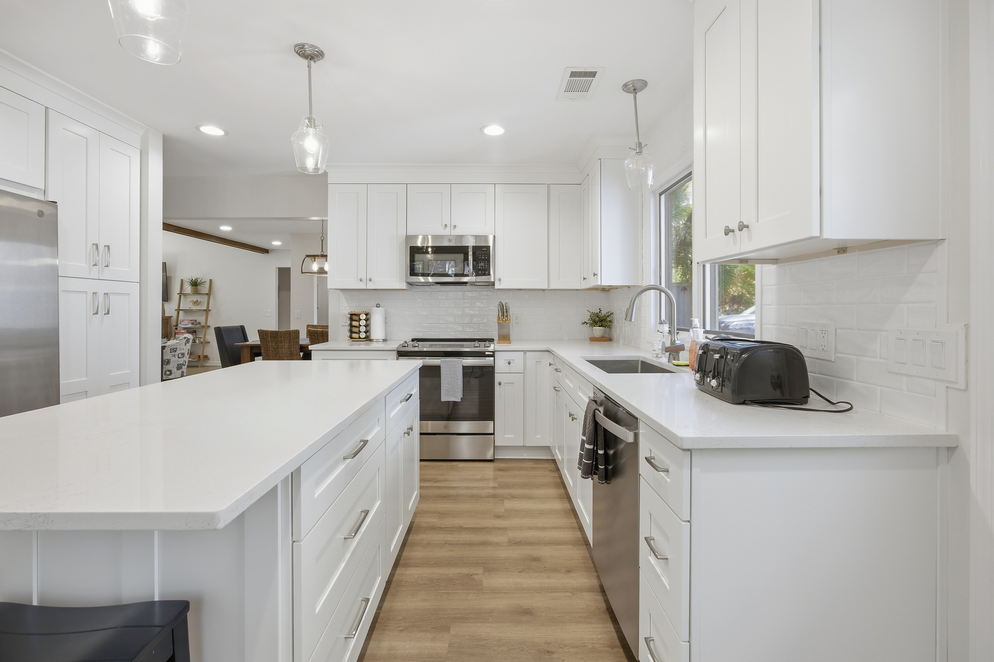 Quartz countertops and snowy cabinetry create a sleek, airy kitchen perfect for slow mornings. Glass pendant lights glow above the wide island, while hardwood floors pair with stainless accents to offer a clean and modern culinary space.