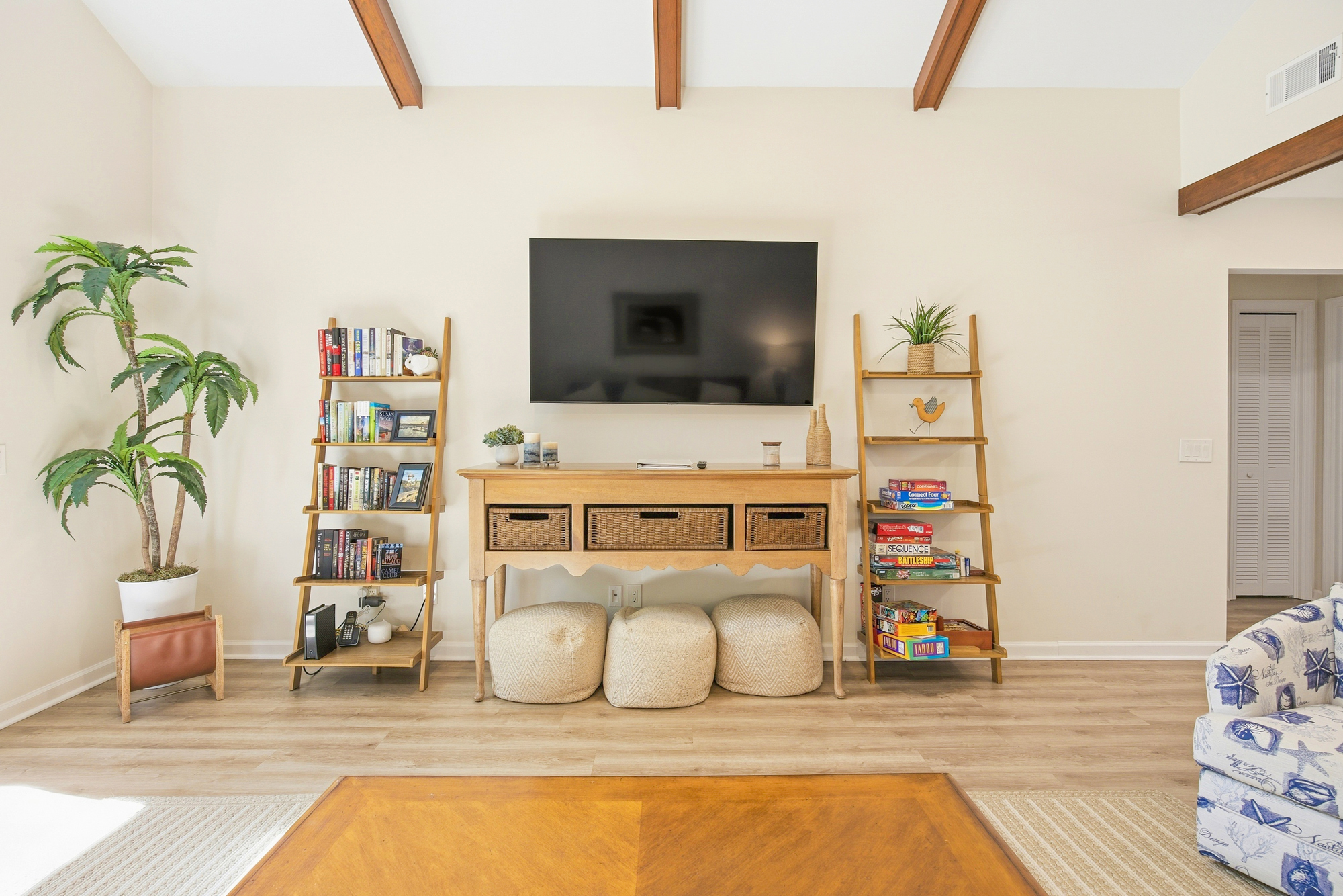 Warm timber beams stretch across the ceiling, framing a cozy entertainment nook filled with board games and reading material. Creamy textures and soft greenery create a calm atmosphere perfect for a quiet afternoon in the lounge.