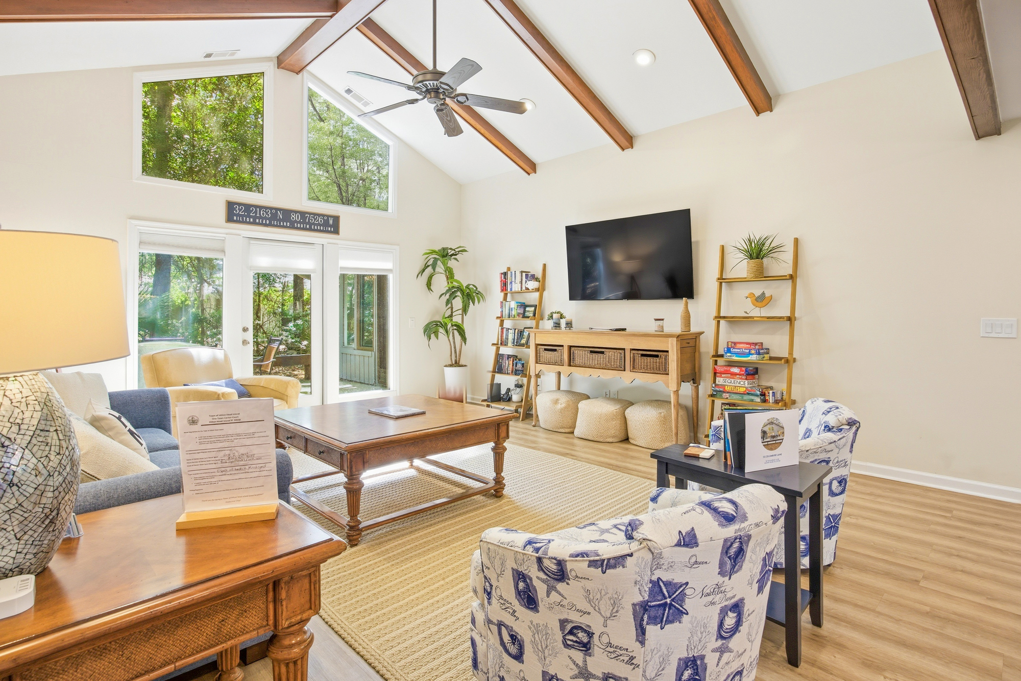 Natural wood beams draw the eye upward in this airy living room, where towering windows frame a verdant forest view. Soft coastal patterns and honey-toned furniture create a grounded, serene space for quiet mornings spent with a book.
