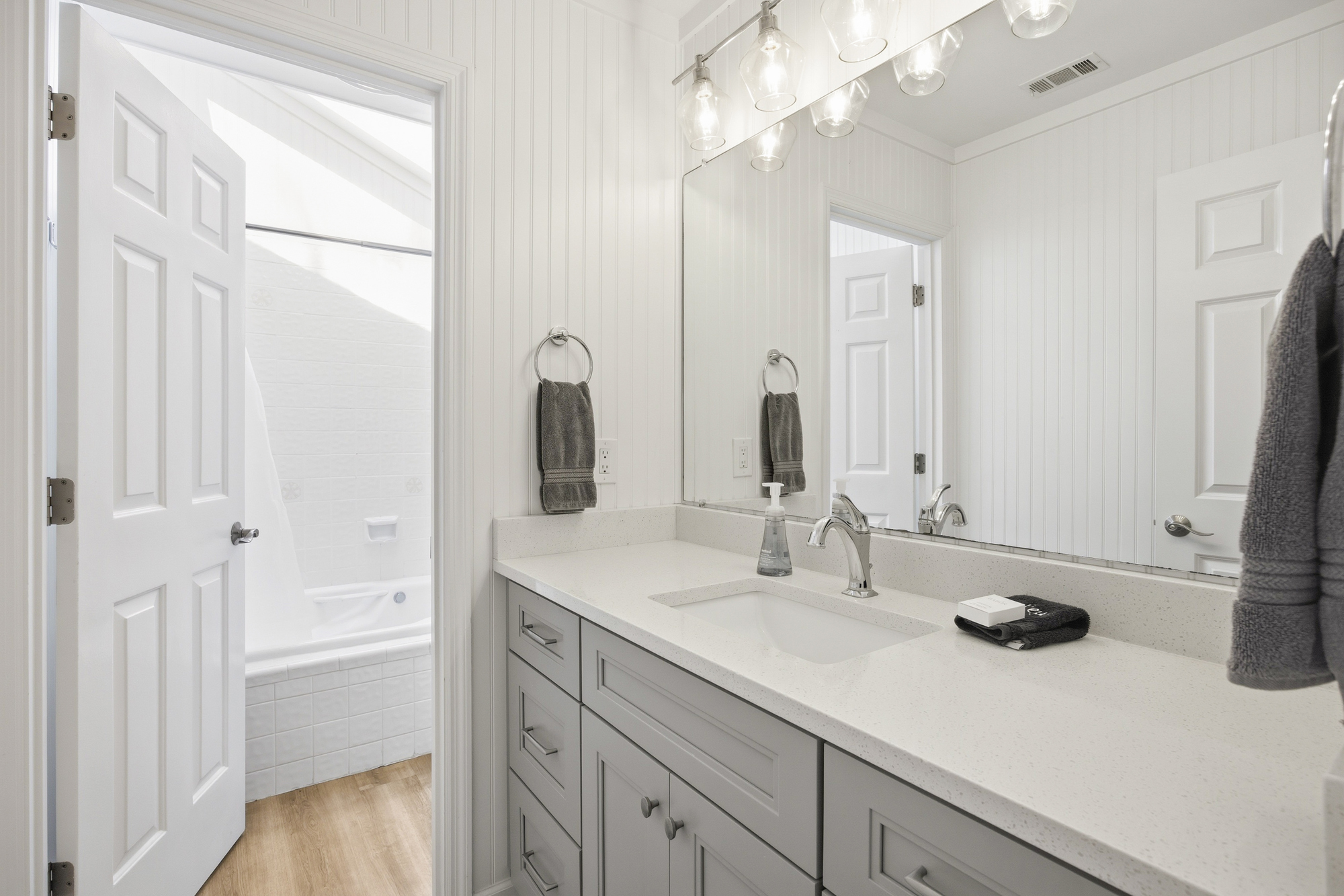 Luminous glass pendants glow over a speckled stone vanity and shiplap walls, creating a serene corner for morning routines. The airy bathroom features soft gray cabinetry and a glimpse into the pristine tiled tub area.