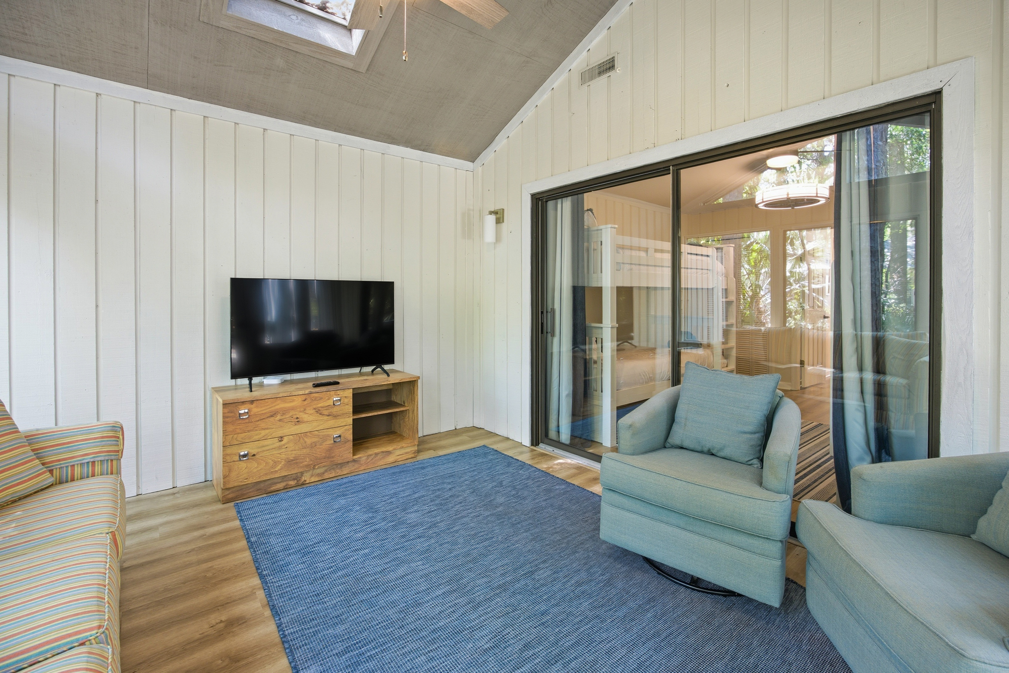 Plush teal armchairs and a textured cobalt rug create a coastal lounge perfect for movie nights. Bright sky-lights and paneled walls bounce natural glow across the warm wood floors, while glass doors seamlessly connect this airy nook to the bedroom.