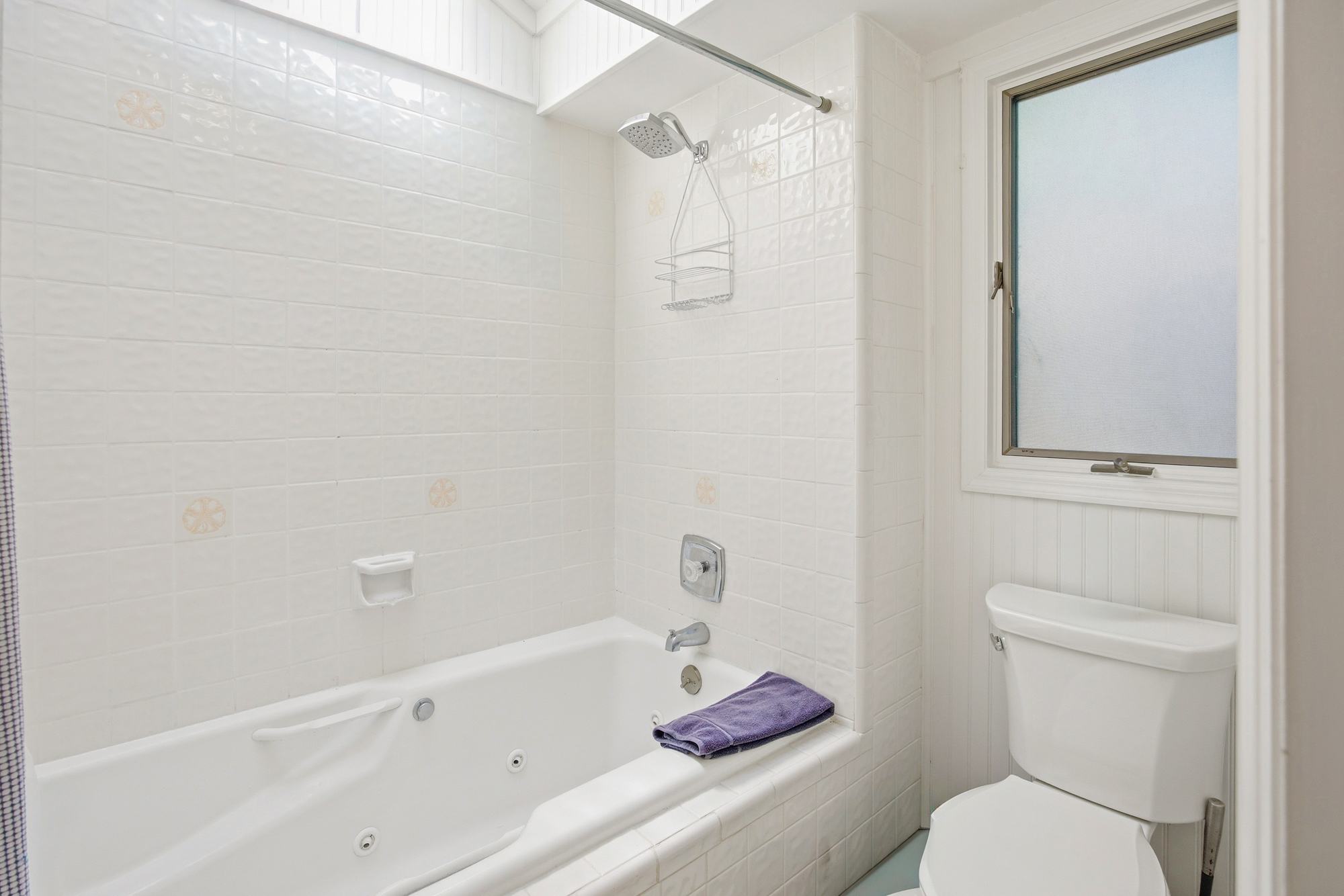 Glistening white tiles and a deep soaking tub create a serene corner for rejuvenation. Soft morning rays filter through the window, highlighting the clean lines and airy atmosphere of this peaceful bathroom.