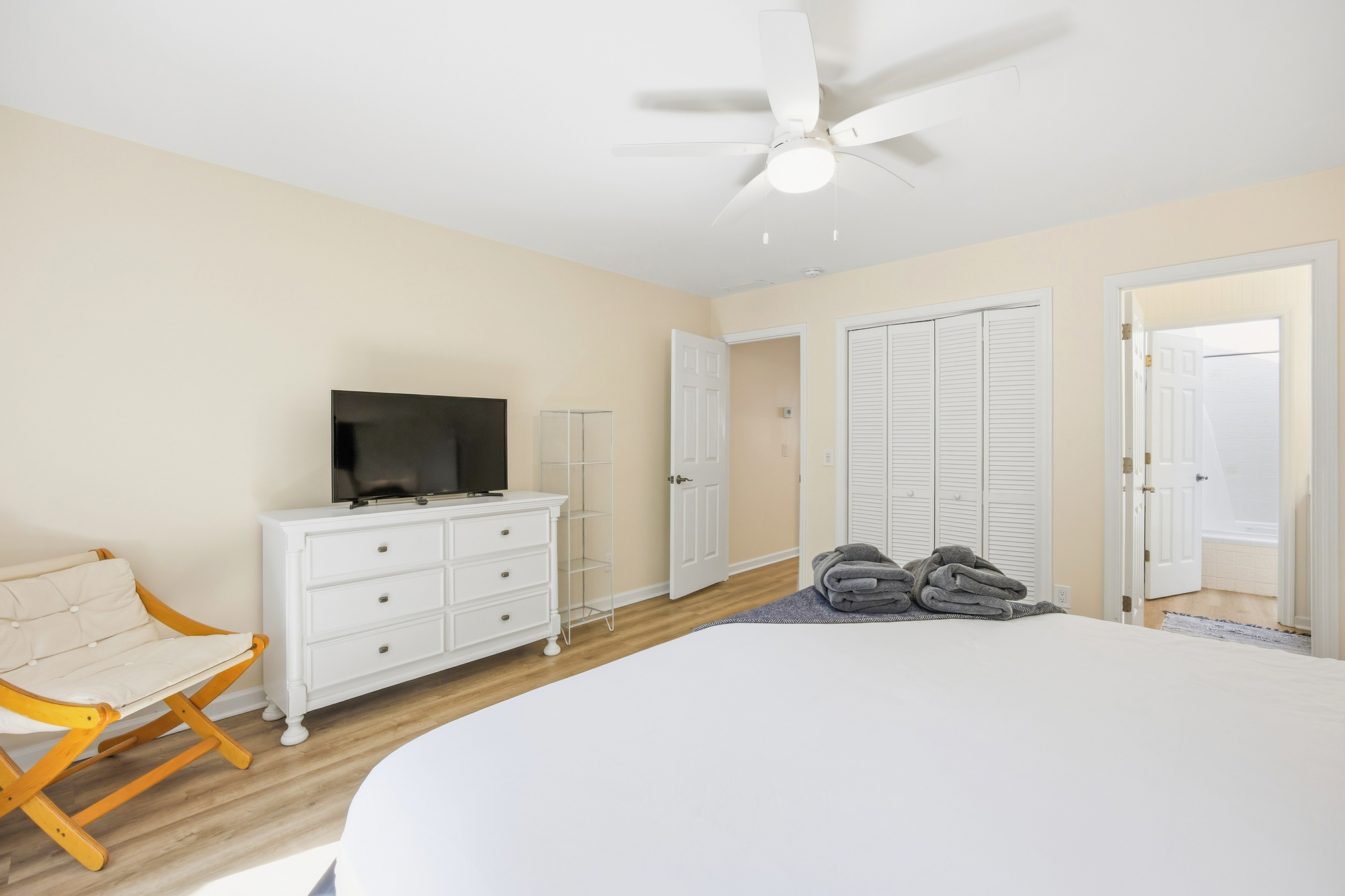 Creamy walls and honey-toned flooring create a serene backdrop for this airy bedroom. A classic white dresser and louvred closet doors add a timeless touch, while the soft linens and natural wood chair suggest a peaceful spot for rest.