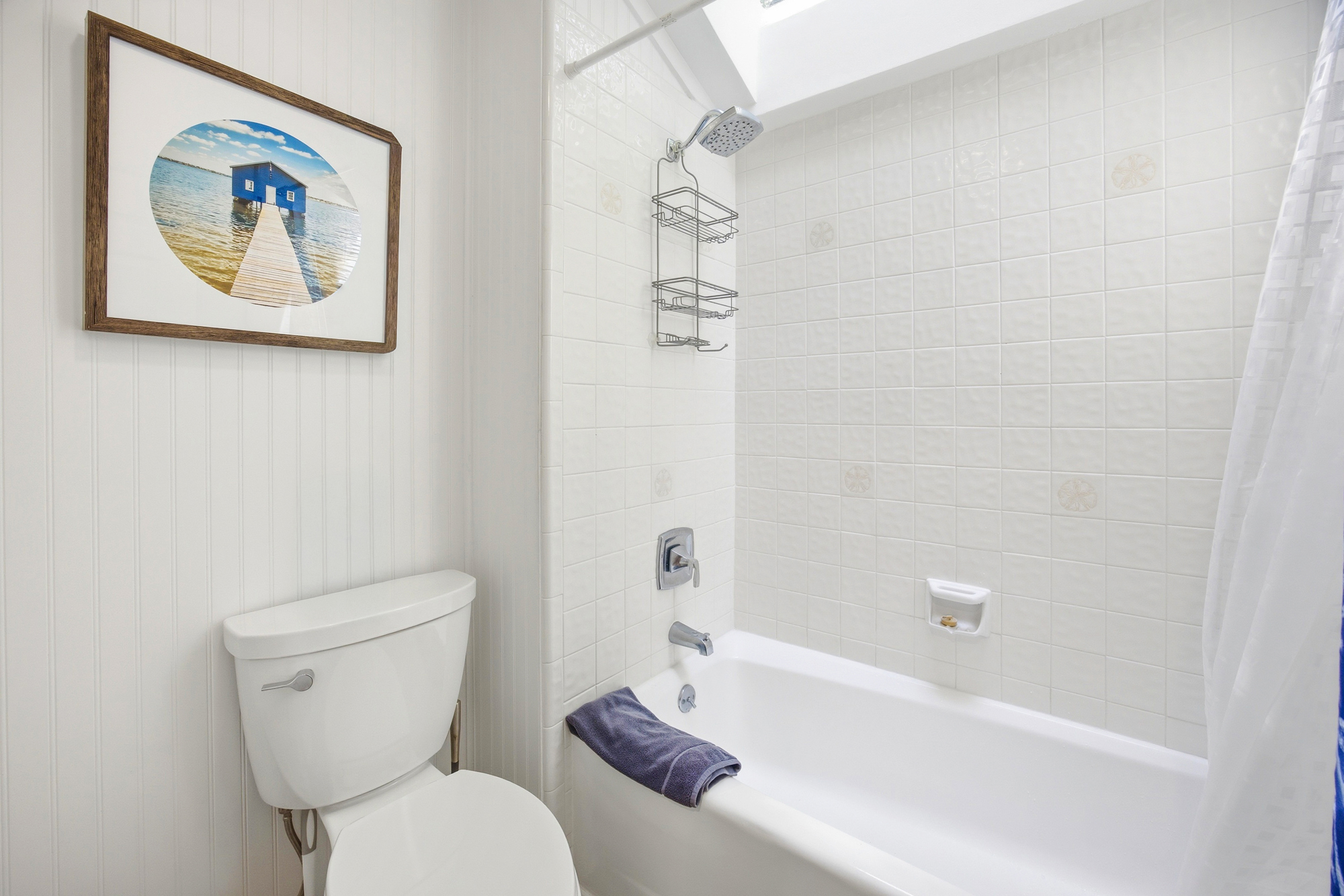 White tiles and beadboard walls create a bright, airy backdrop for a refreshing morning rinse. Natural glow pours through the skylight, illuminating the deep soaking tub and calming coastal art for a serene start to your day.
