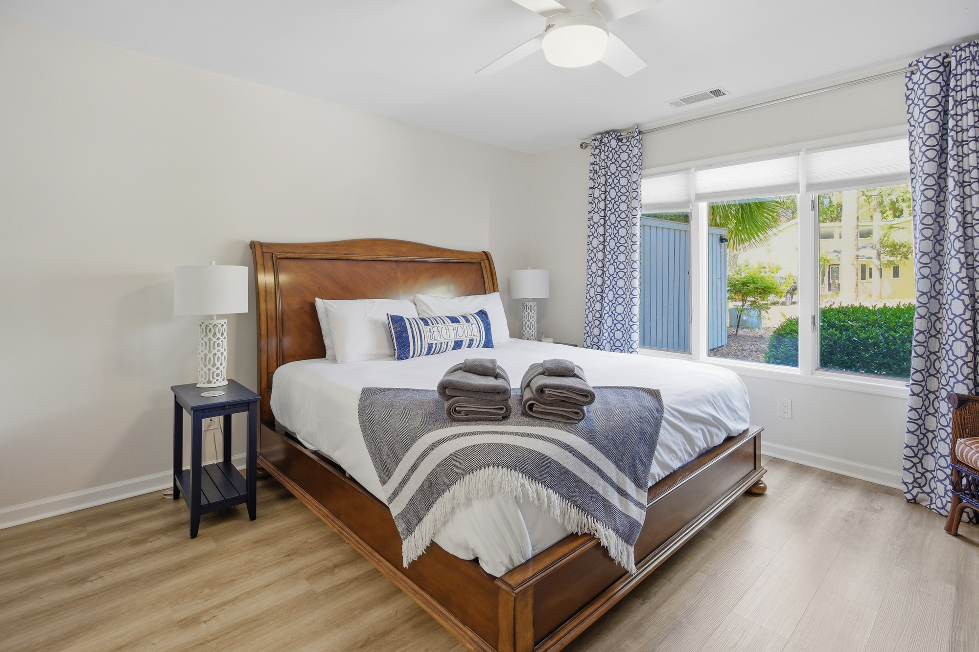 Polished wood and coastal blue tones define this airy bedroom, where a large window invites the morning glow inside. Soft linens and a fringed throw wait on the king bed, creating a calm space for a restful night by the sea.