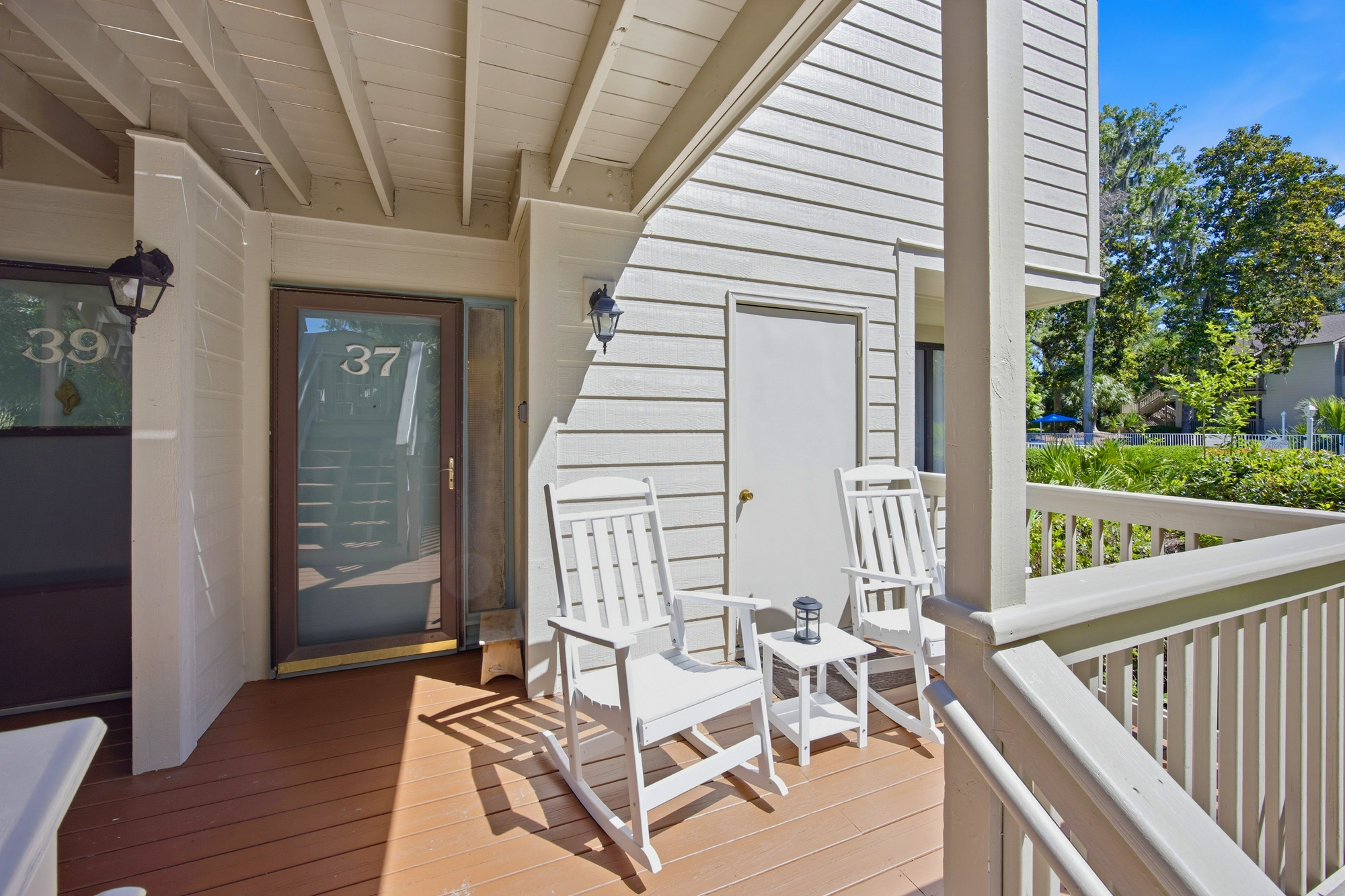 Crisp white rocking chairs sit on a private wooden deck, catching the soft afternoon glow. This quiet corner overlooks lush greenery and offers a peaceful spot to breathe in the fresh air under a clear blue sky.
