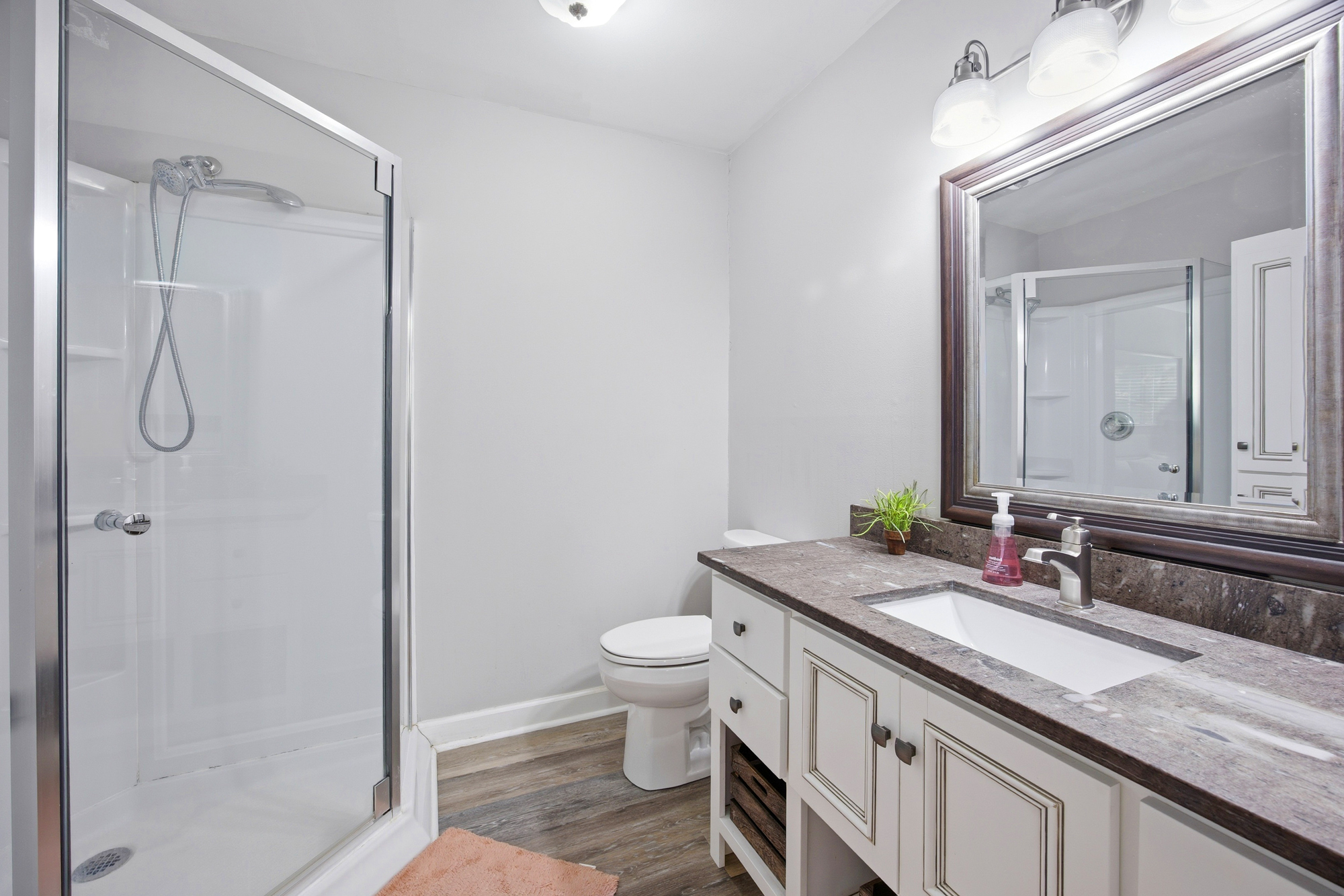 Polished stone counters and bright white cabinetry create a clean, refreshing atmosphere for your morning routine. A spacious glass-enclosed shower and warm wood floors offer a modern, spa-like feel.
