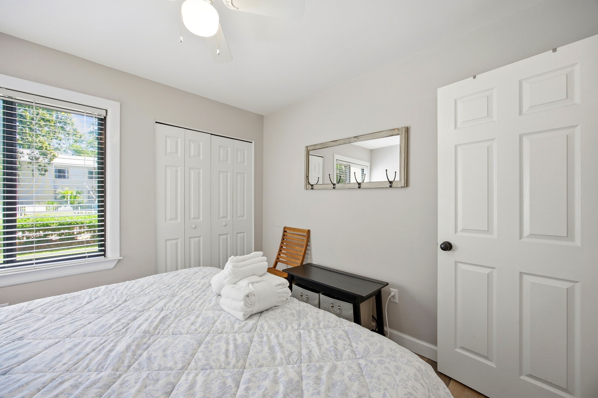 Fluffy white towels wait atop a quilted bed in this calm bedroom, where a leafy view filters through the blinds. Gentle daylight fills the space, bouncing off the clean white surfaces and a rustic wall mirror to create a welcoming atmosphere.