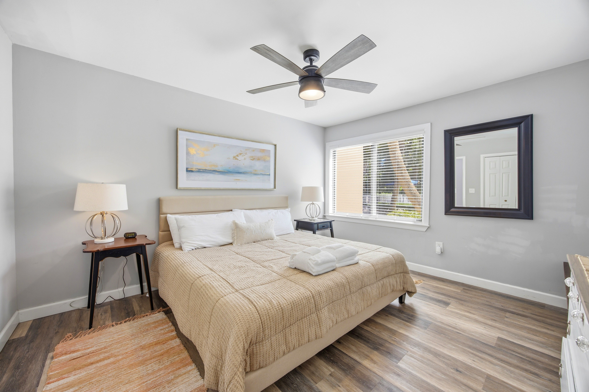 Pale textures and cool gray walls create a calm atmosphere, while shifting daylight fills the room with a gentle glow. A plush bed sits atop rich wood flooring, offering a quiet space to rest and recharge.