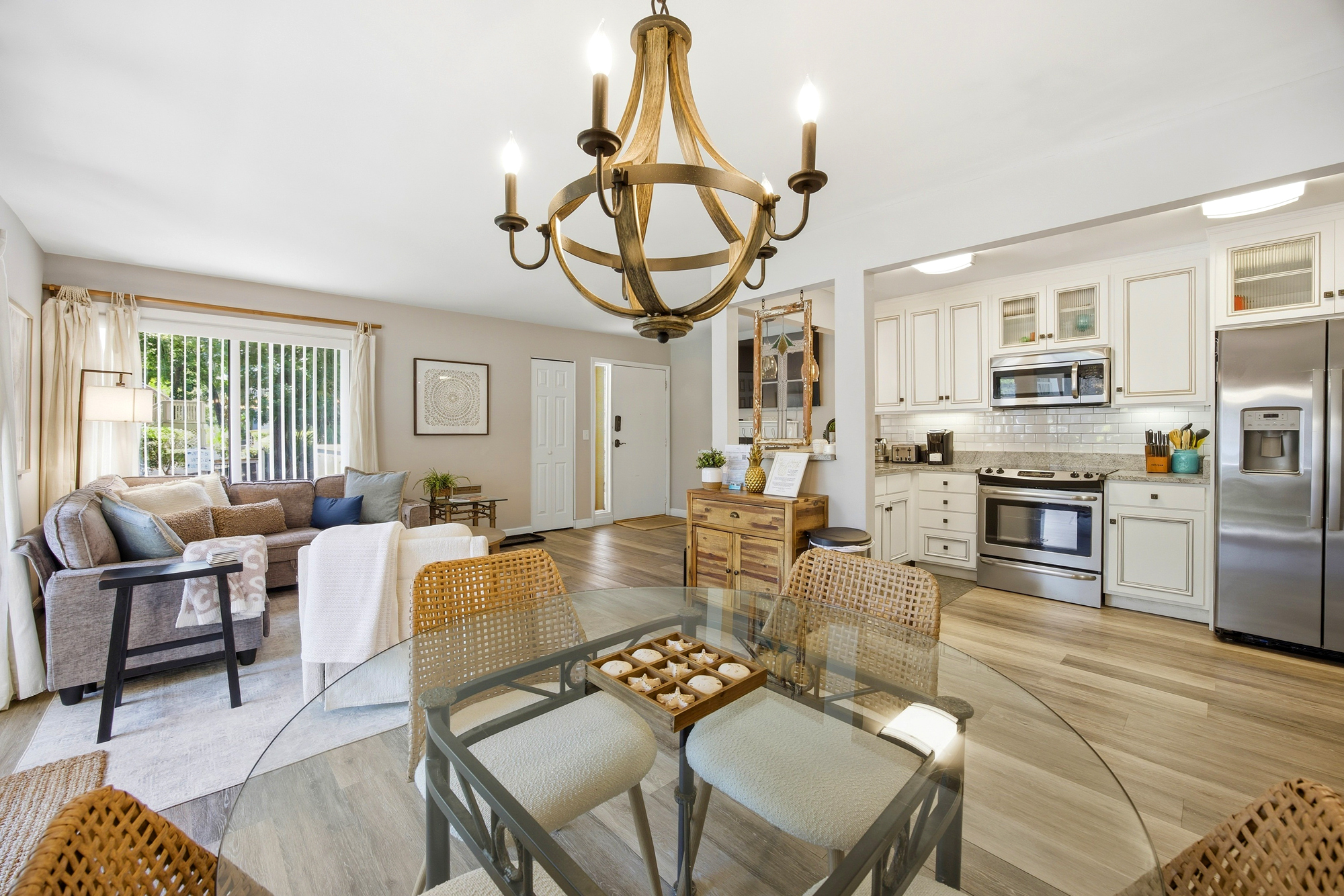 A glass dining table and rattan chairs anchor this airy open floor plan, where white cabinetry meets warm wood accents. Golden light spills across the wide-plank floors, connecting the modern kitchen to a cozy lounge perfect for long conversations.