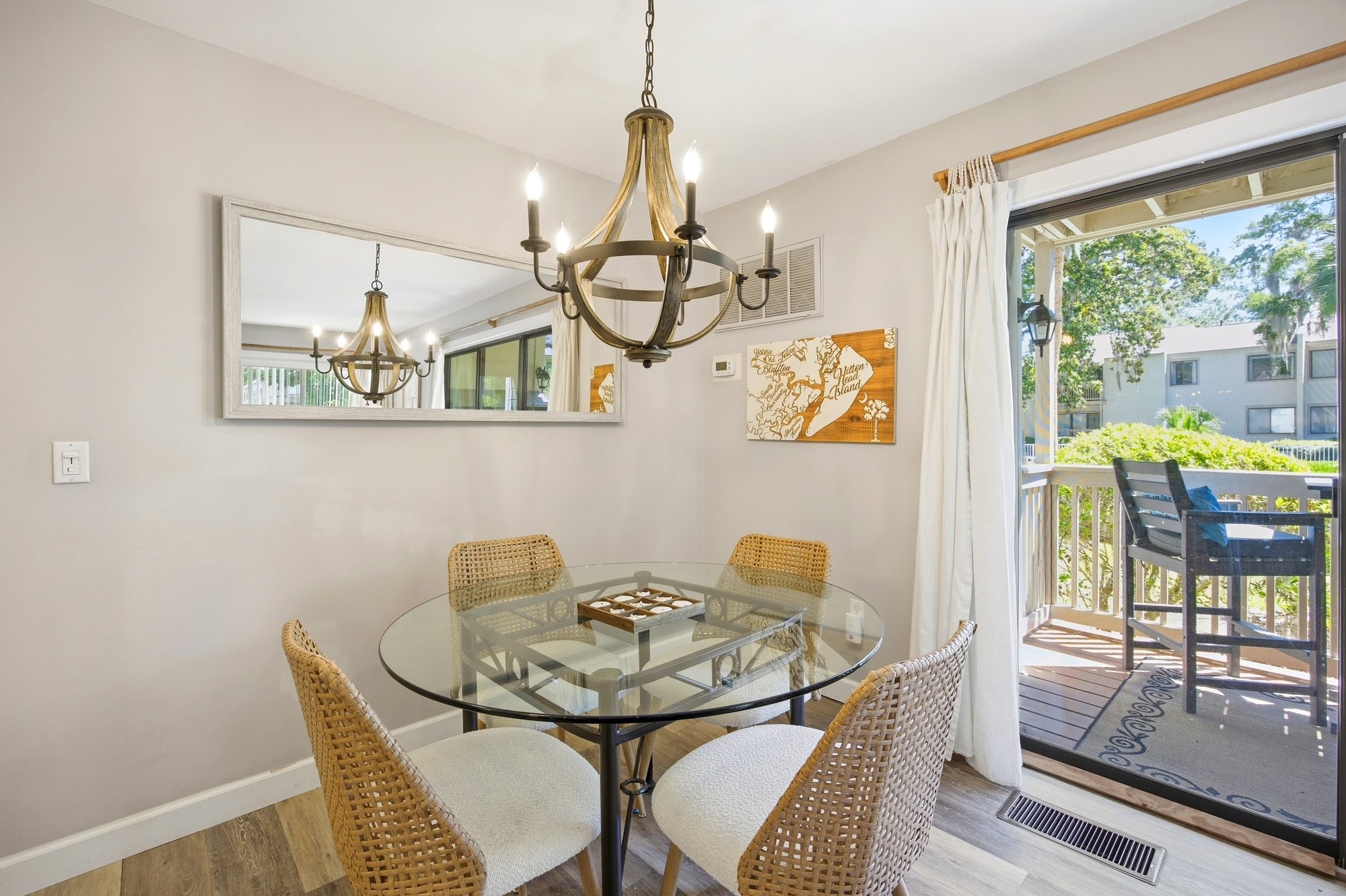 Rattan seating and a glass-topped table create a breezy spot for morning coffee beside the sun-drenched balcony. The rustic chandelier casts a gentle glow over the open dining area, perfect for sharing meals and planning island adventures.