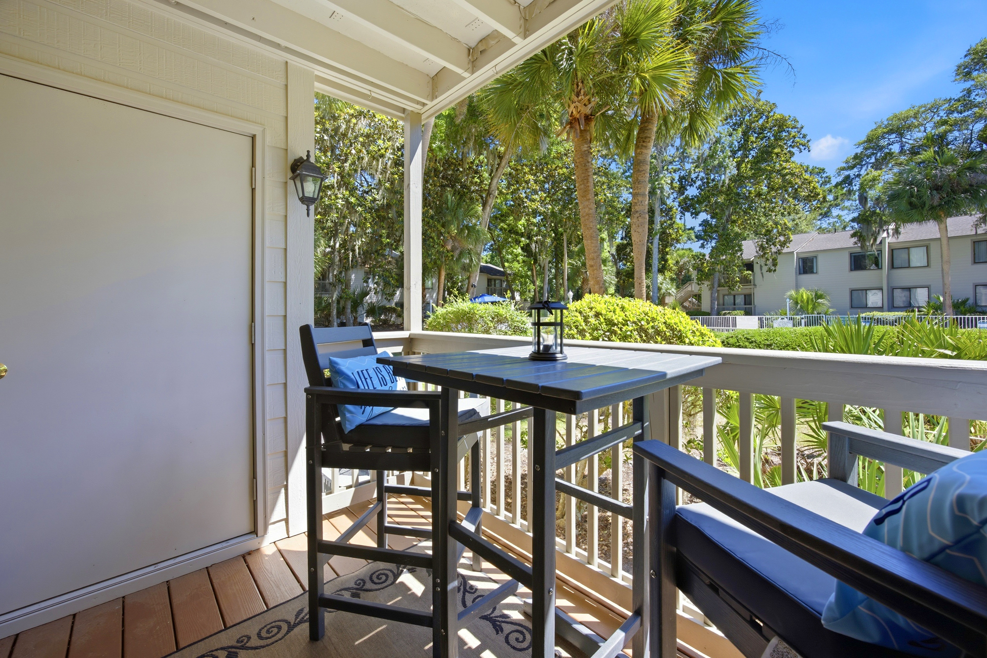Palm trees sway beyond this covered balcony, where a high-top table sits ready for coffee. Golden rays illuminate the timber floorboards, creating a quiet spot to watch the world go by.