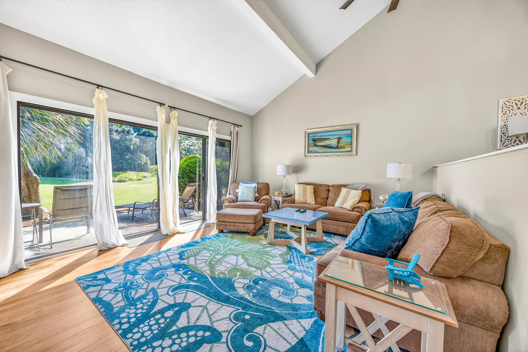 Azure waves dance across the rug in this airy living room, where vaulted ceilings and glass doors invite the garden inside. Plush seating in earthy tones offers a cozy spot to gather as the midday brightness fills every corner of the space.