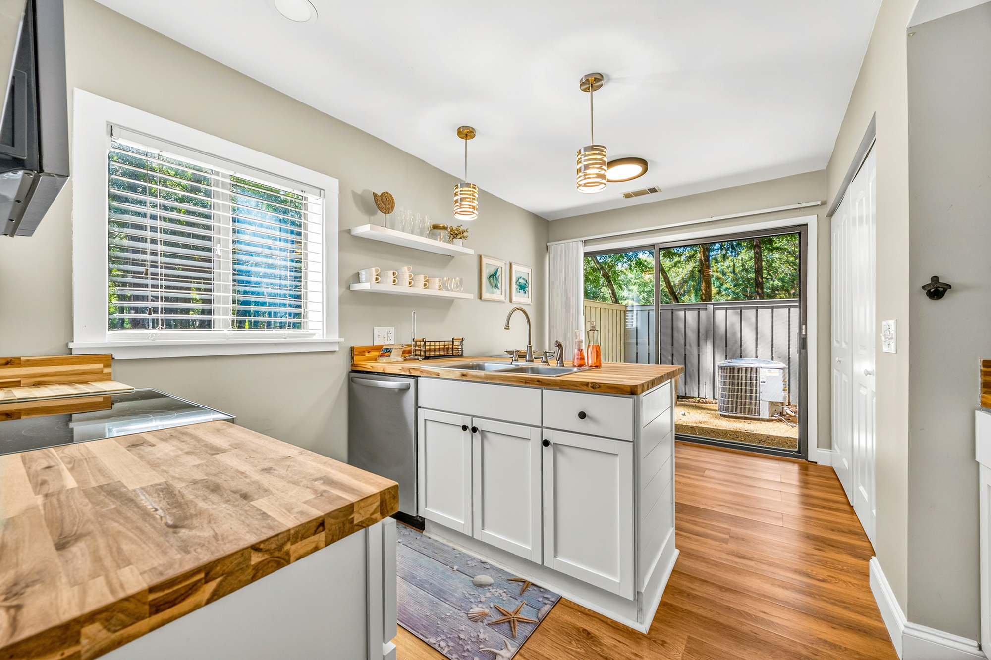 Honey-toned butcher block counters and warm wood flooring create a grounded, organic feel in this sun-drenched kitchen. Modern gold pendants glow above the island, leading your eye toward the lush greenery visible through the sliding glass doors.