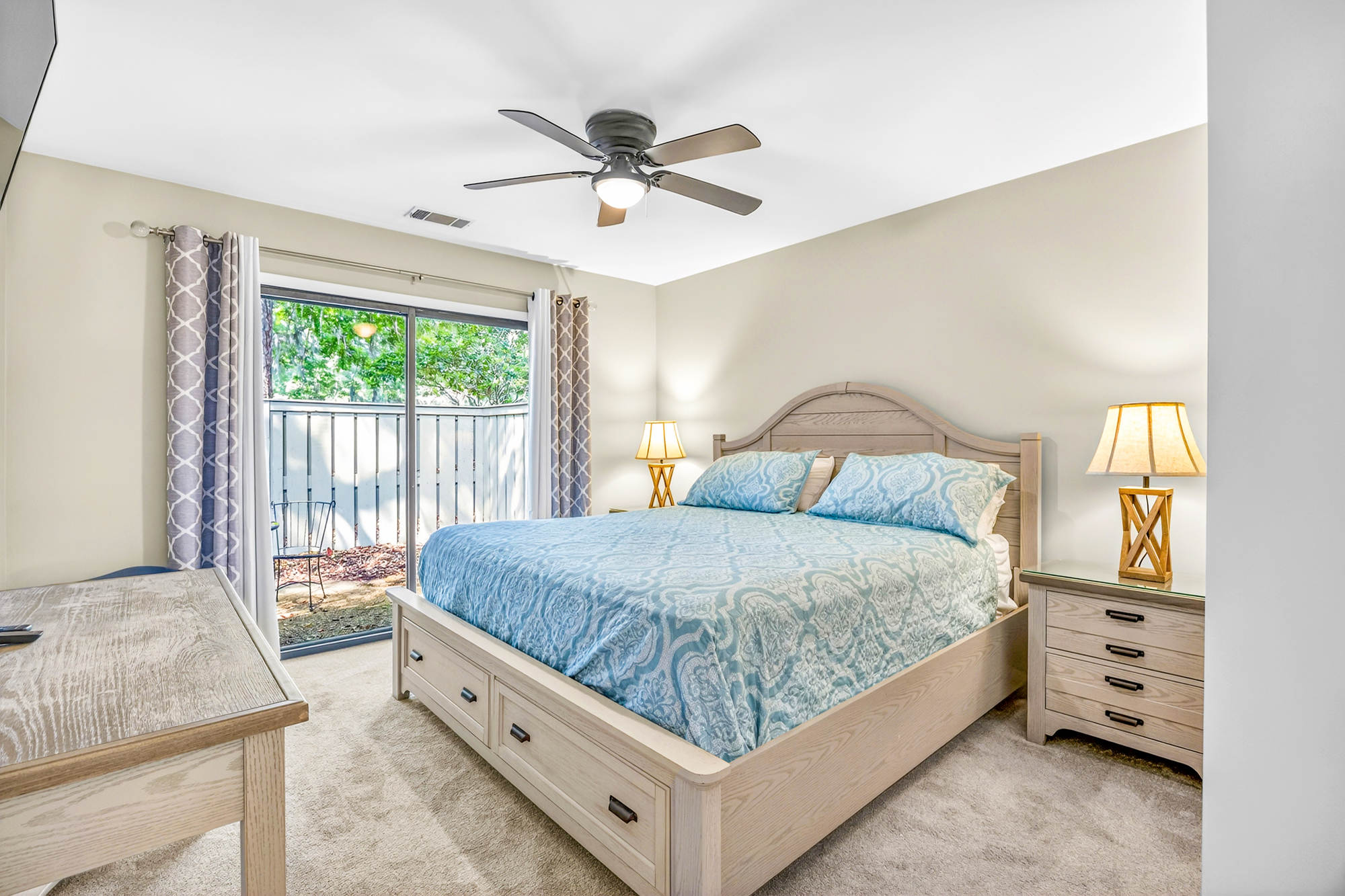 Soft teal patterns and pale wood grains create a serene atmosphere in this airy bedroom. Sliding glass doors pull in the lush garden views, while gold lamp light adds a cozy glow to the quiet, plush surroundings.