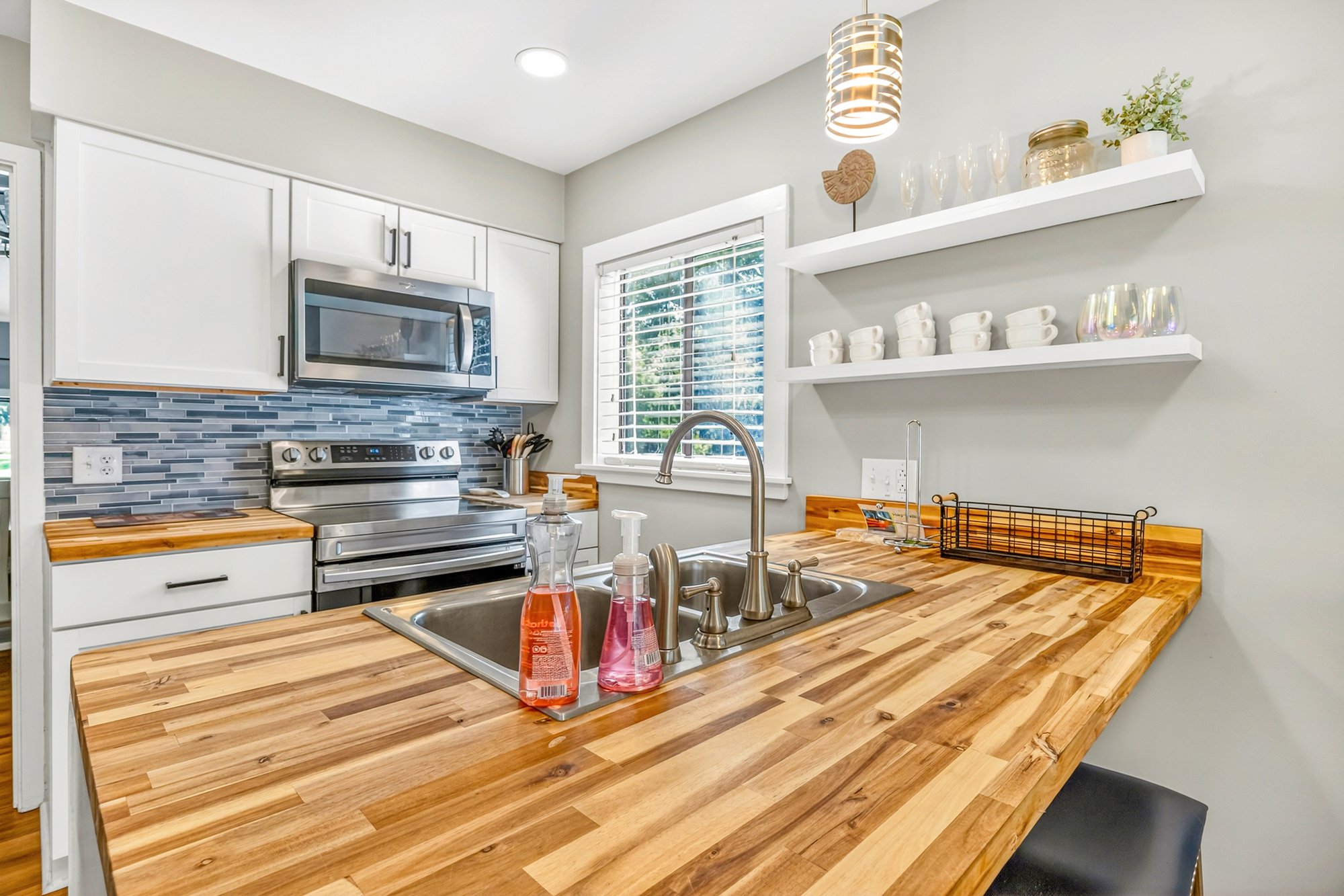 Polished butcher block counters bring a rich, organic warmth to this modern kitchen. Soft daylight filters through the window, illuminating the textured blue backsplash and crisp white floating shelves for a bright, airy feel.