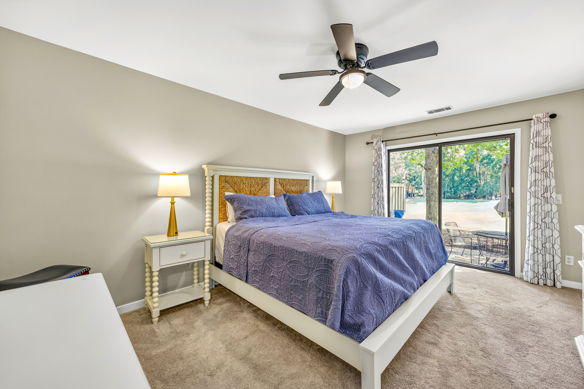 Woven textures and deep sapphire linens create a peaceful sanctuary for restful nights. Sliding glass doors pull the lush greenery inside, while golden lamp light adds a cozy, amber glow to the plush carpet and clean, white furnishings.