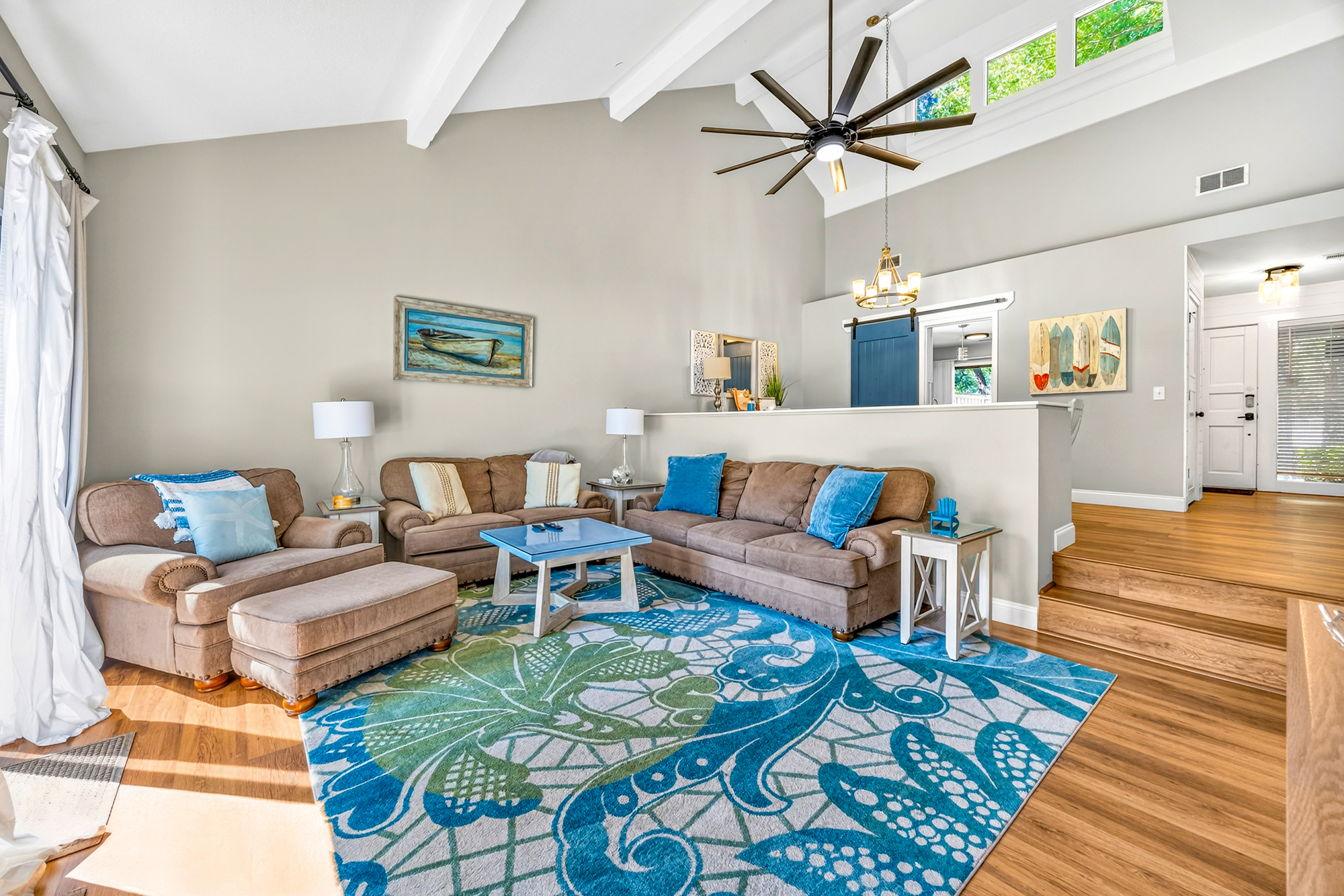 Vaulted ceilings and clerestory windows flood this coastal living room with a bright afternoon glow. Plush seating anchored by a vibrant teal rug offers a cheerful space to gather on warm oak floors.