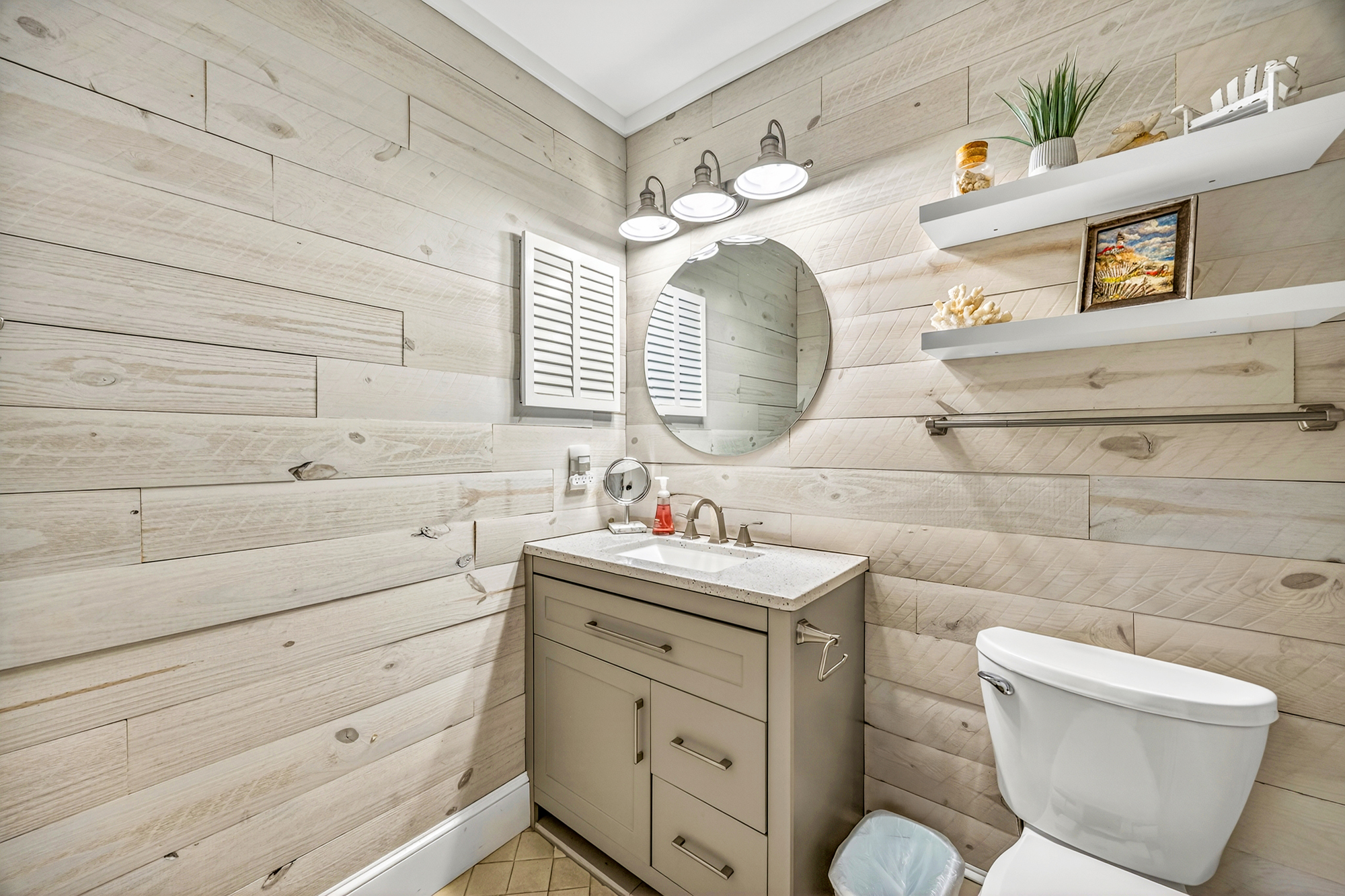 Whitewashed timber walls bring a coastal serenity to this bright bathroom. A circular mirror and industrial lighting provide a modern touch, while maritime accents on floating shelves create a charming, airy atmosphere for your morning routine.