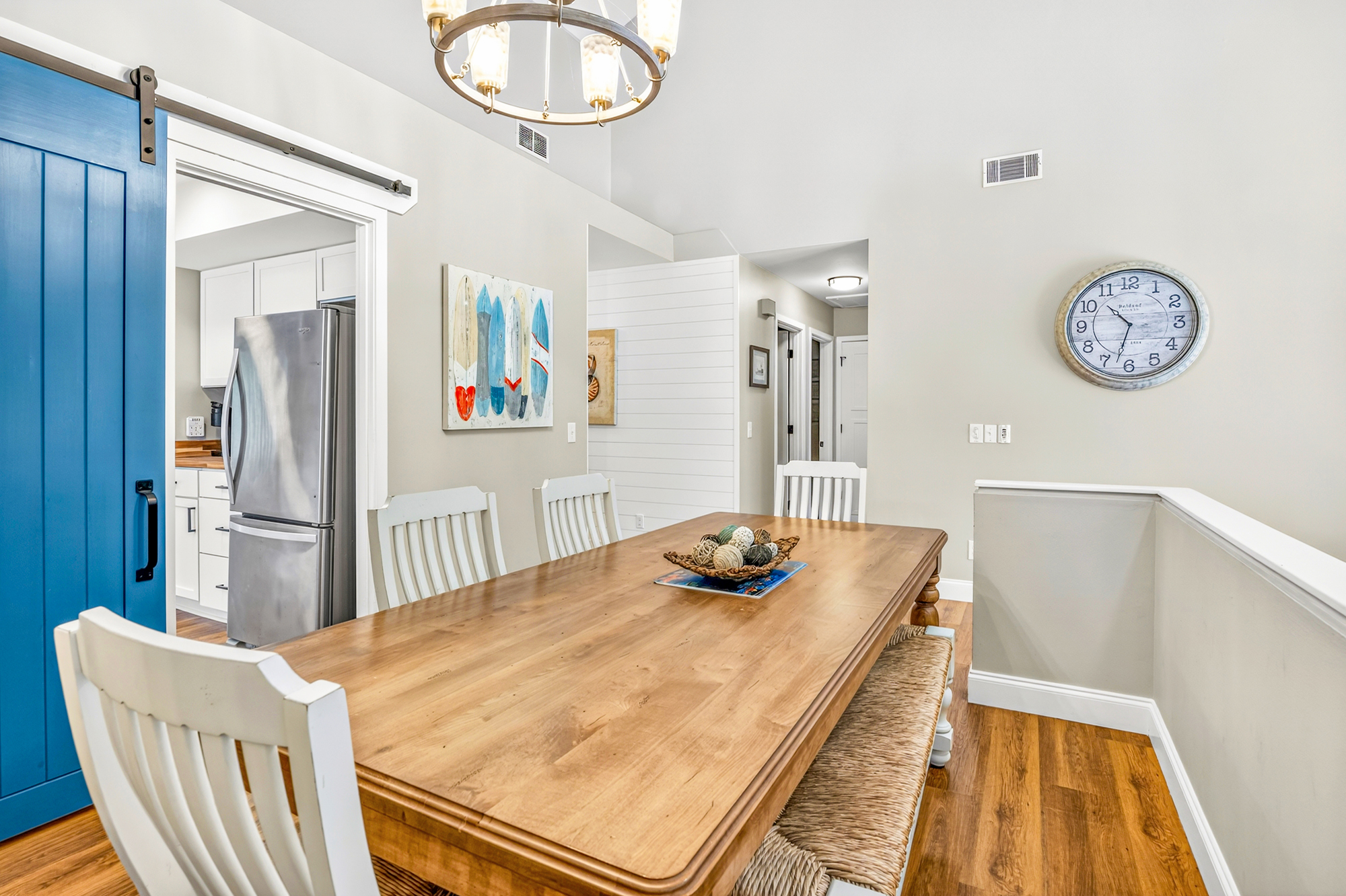 Oak grain and white slatted chairs create a coastal dining nook, anchored by a bold blue barn door and a nautical chandelier. Seagrass seating and surfboard art bring a beachy soul to this airy, open gathering place.