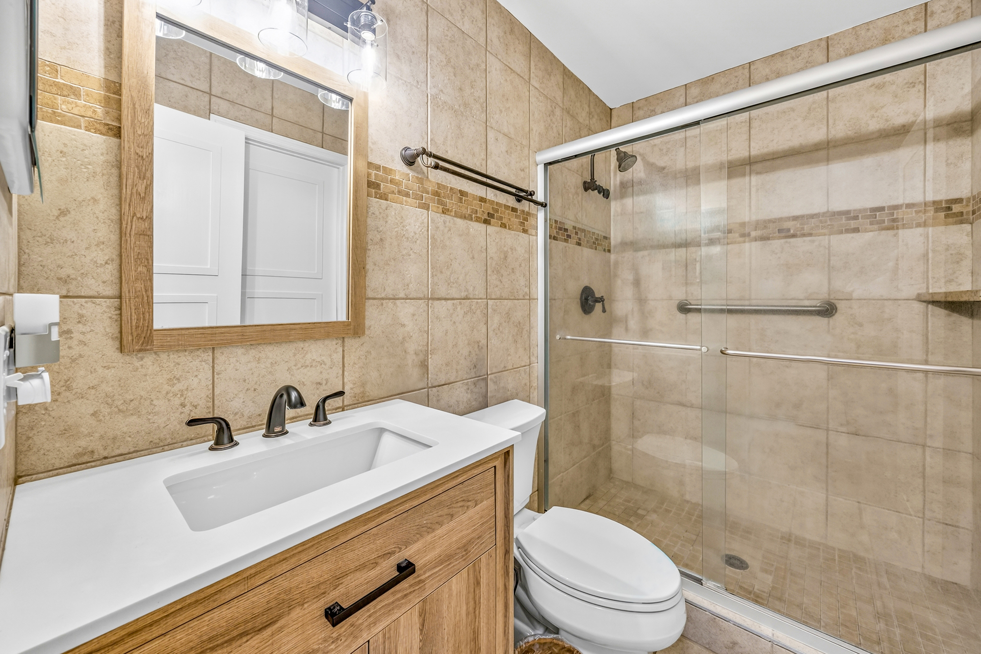 Earth-toned tiles and a warm wood vanity create a spa-like atmosphere in this bright, clean bathroom. A spacious glass-enclosed shower and modern fixtures offer a refreshing space to begin your day.