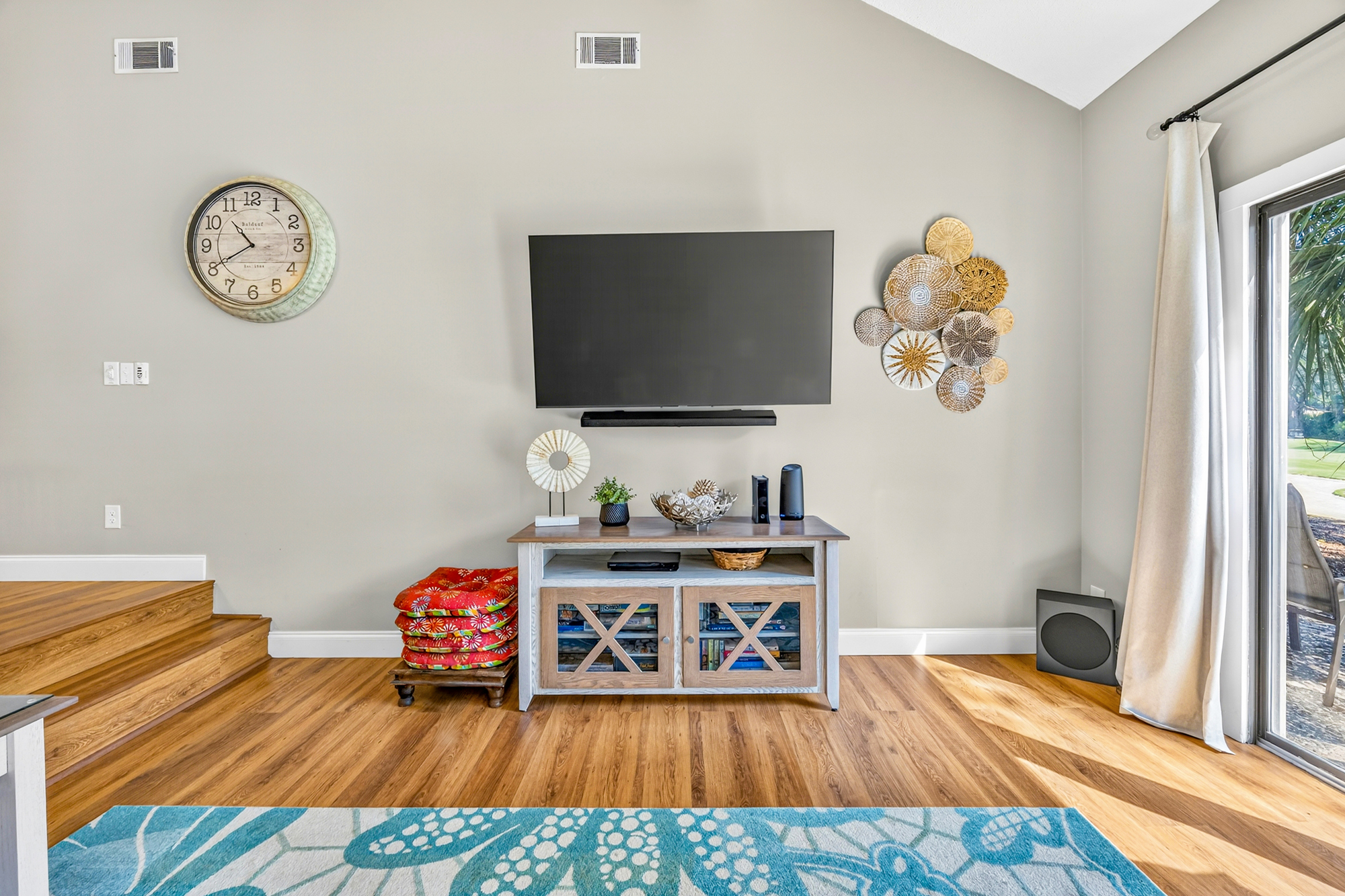 Warm oak floors glow under the afternoon radiance streaming through glass doors, creating a cozy media nook. Textured wall art and beachy tones balance the sleek entertainment setup for a calm evening in.