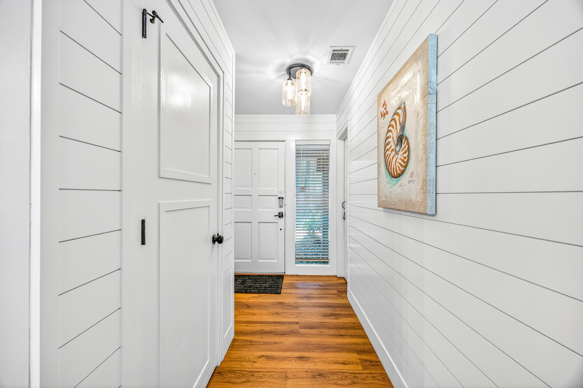 Horizontal shiplap walls lead the eye toward a bright entryway, where nautical art and amber wood flooring create a coastal greet. Modern glass pendants cast a gentle luster over this clean, airy hallway.