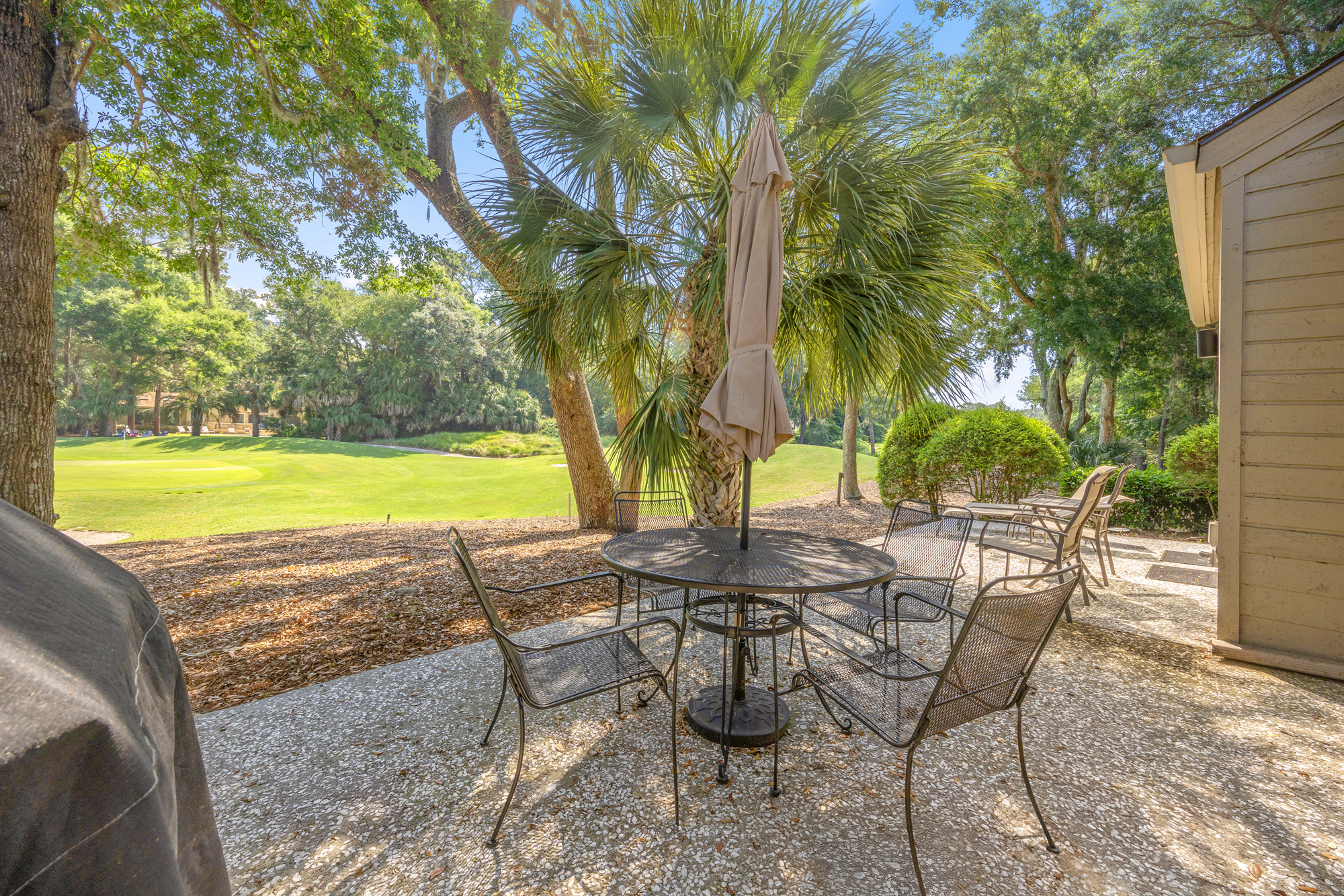 Wrought iron seating sits under the shade of a towering palm, offering a quiet spot to survey the rolling green fairways. Bright afternoon rays filter through the mossy oaks, illuminating this stone patio for a peaceful coffee or sunset meal.