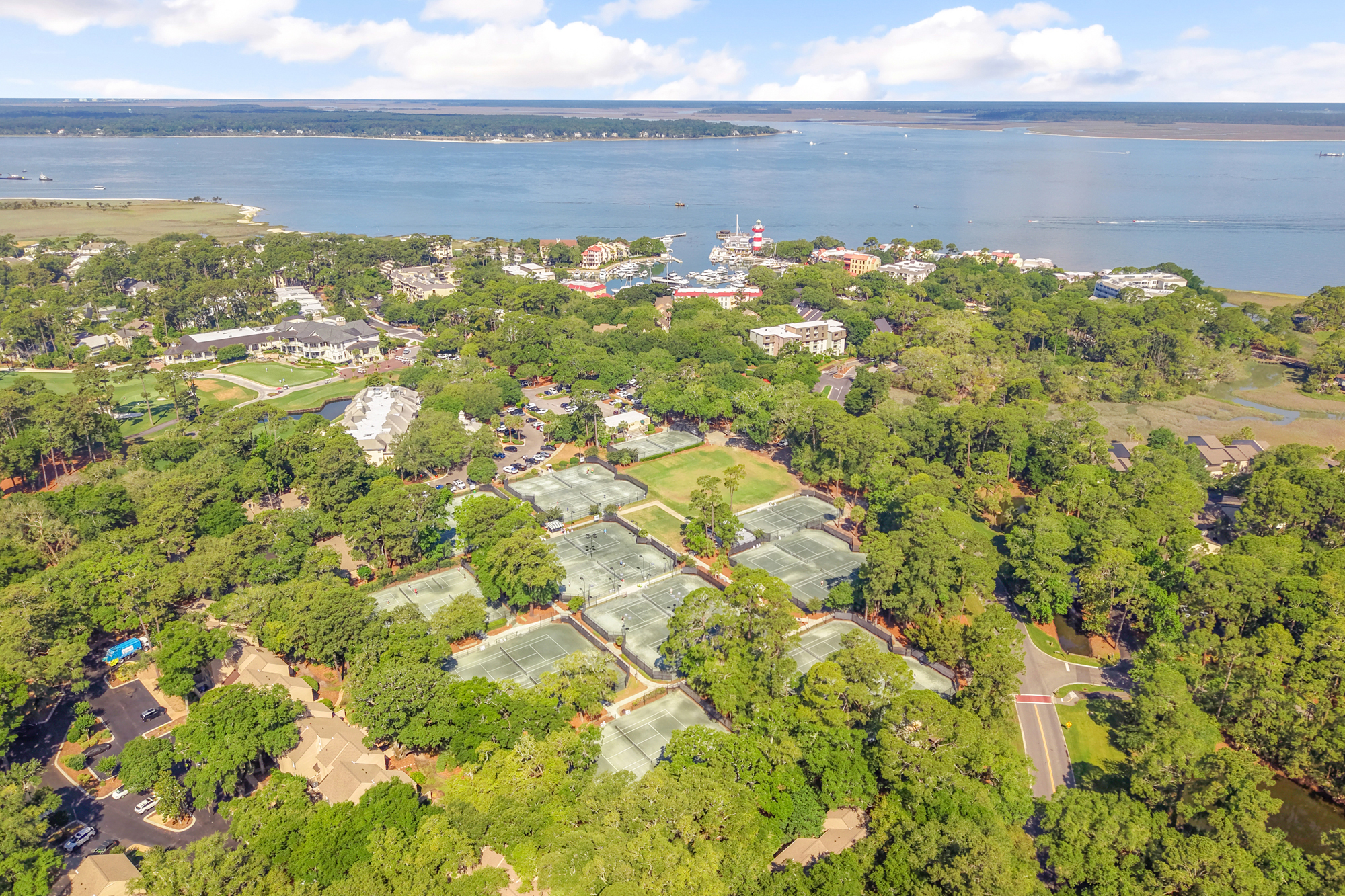 Emerald canopies of ancient trees frame a sprawling collection of tennis courts and coastal villas. Shimmering blue waters meet the horizon, offering a scenic backdrop for active afternoons and peaceful sunset strolls by the marina.