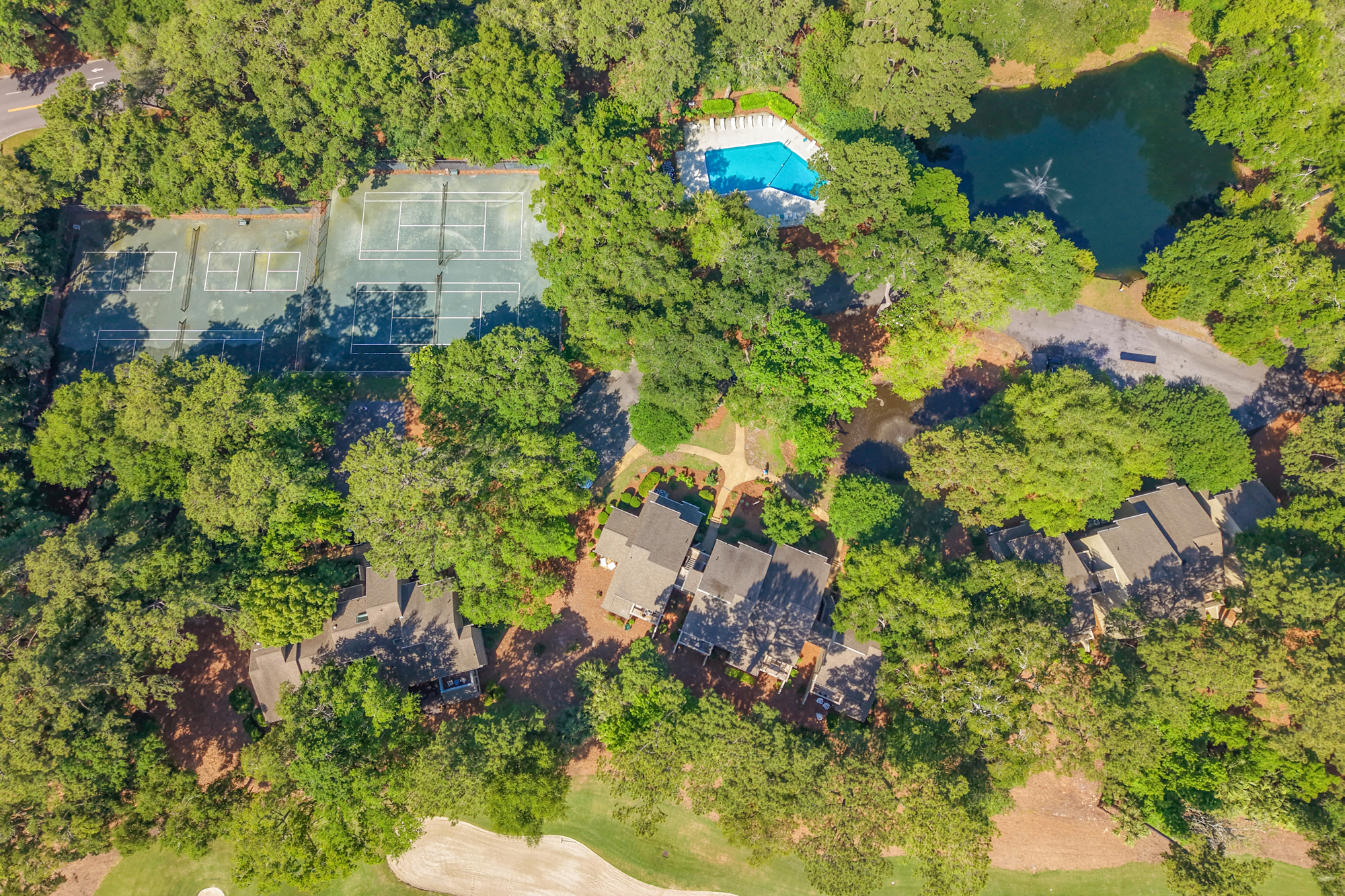 Leafy canopies shade this aerial view of our community, where a sparkling turquoise pool and quiet tennis courts sit just steps from your door. Gentle fountains ripple the dark water of the nearby pond, offering a calm backdrop for your stay.