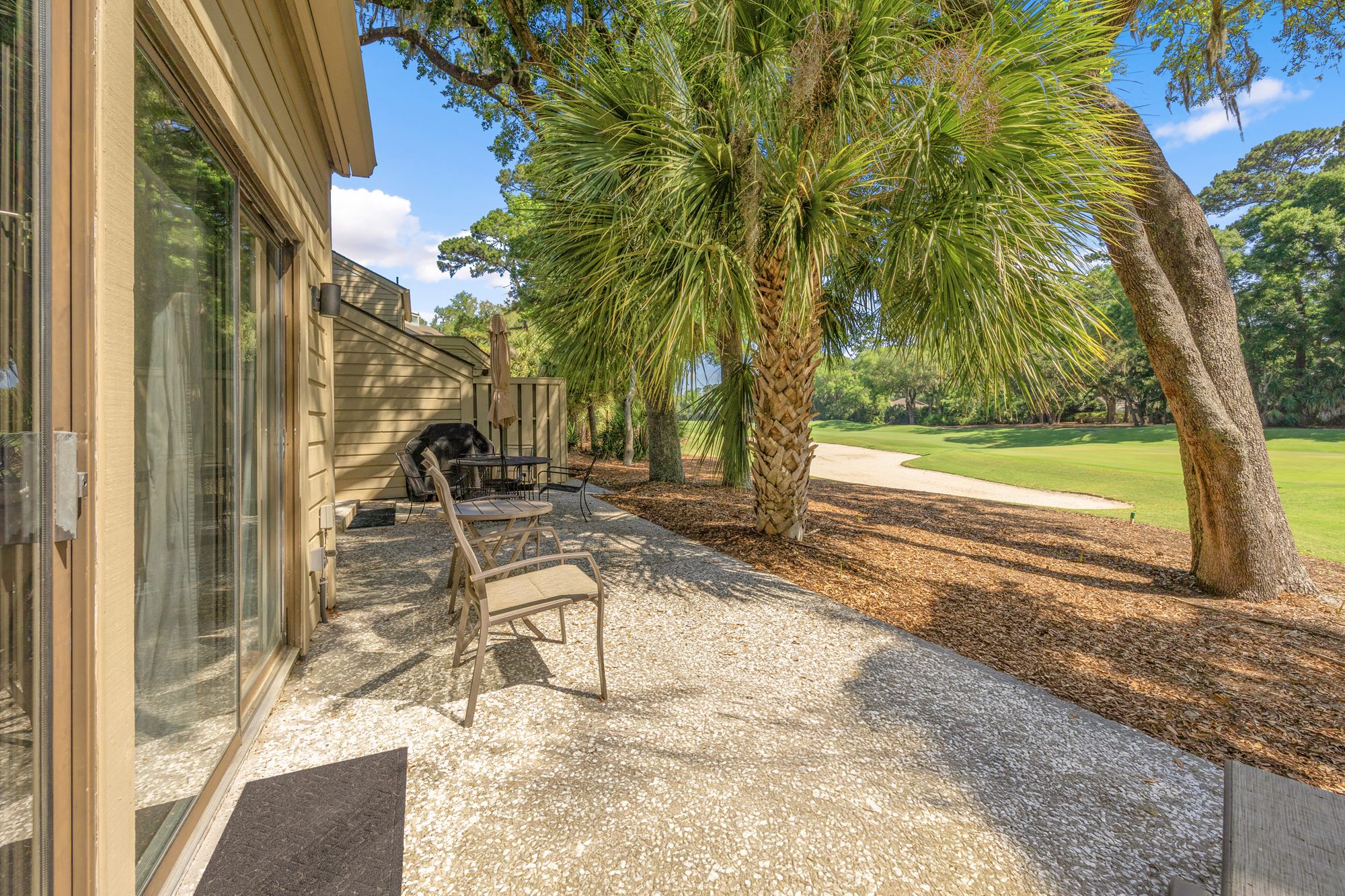 Sun-drenched patio tiles meet the shade of a towering palm, creating a peaceful corner for your morning coffee. This quiet outdoor terrace looks out over a sweeping green fairway framed by ancient oaks and shimmering afternoon light.