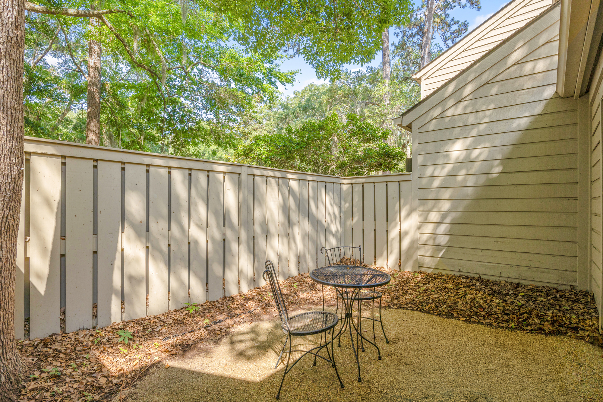 Iron garden chairs sit in a quiet corner, where golden afternoon rays filter through the canopy to dance across the gravel floor. This secluded outdoor nook offers a peaceful spot for coffee among the rustling leaves and tall trees.