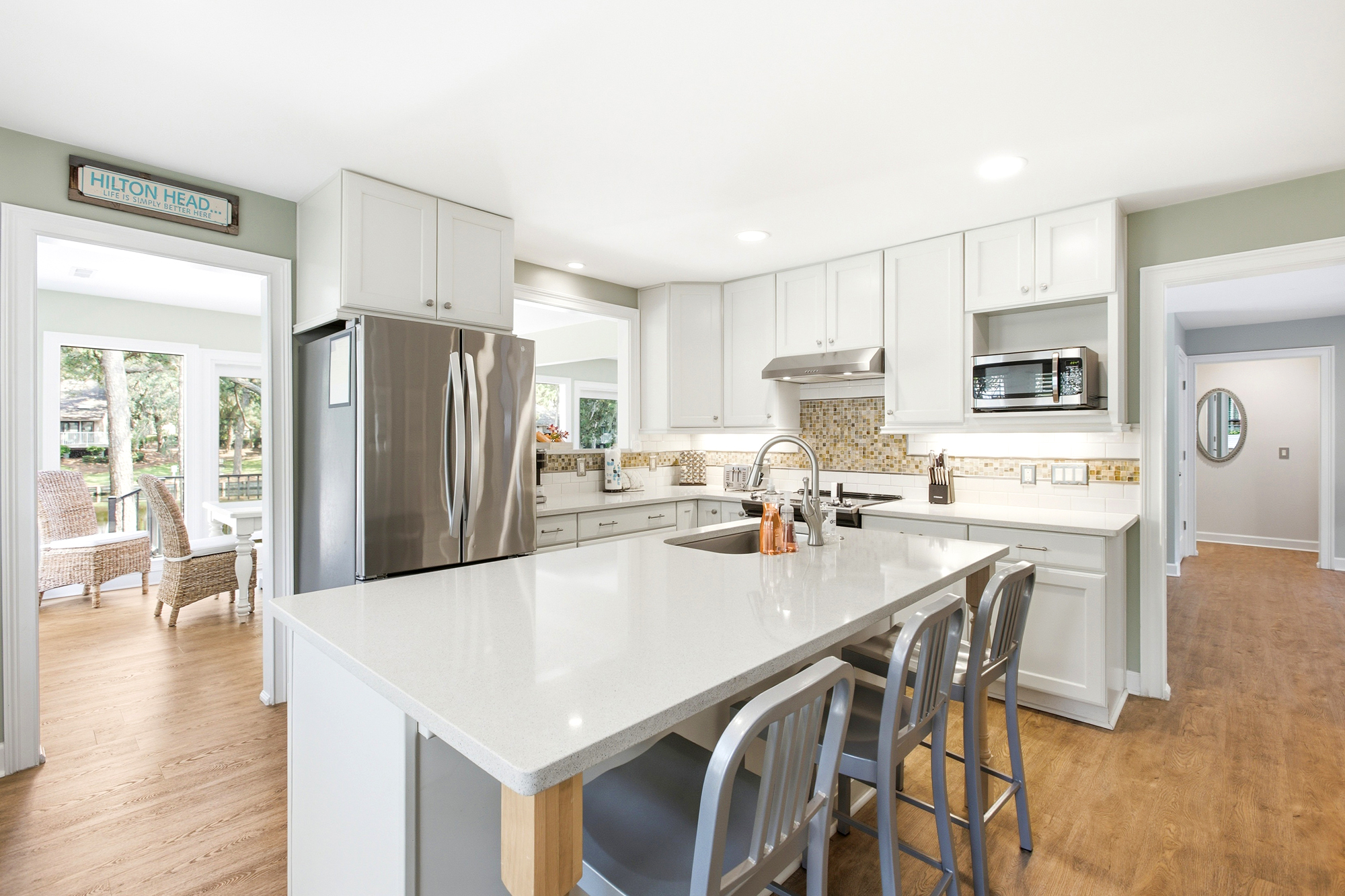 Crisp white cabinetry and cool stone countertops create a bright, airy hub for morning coffee and shared meals. Soft light pours across the warm wood floors, highlighting a modern kitchen designed for effortless hosting and culinary ease.
