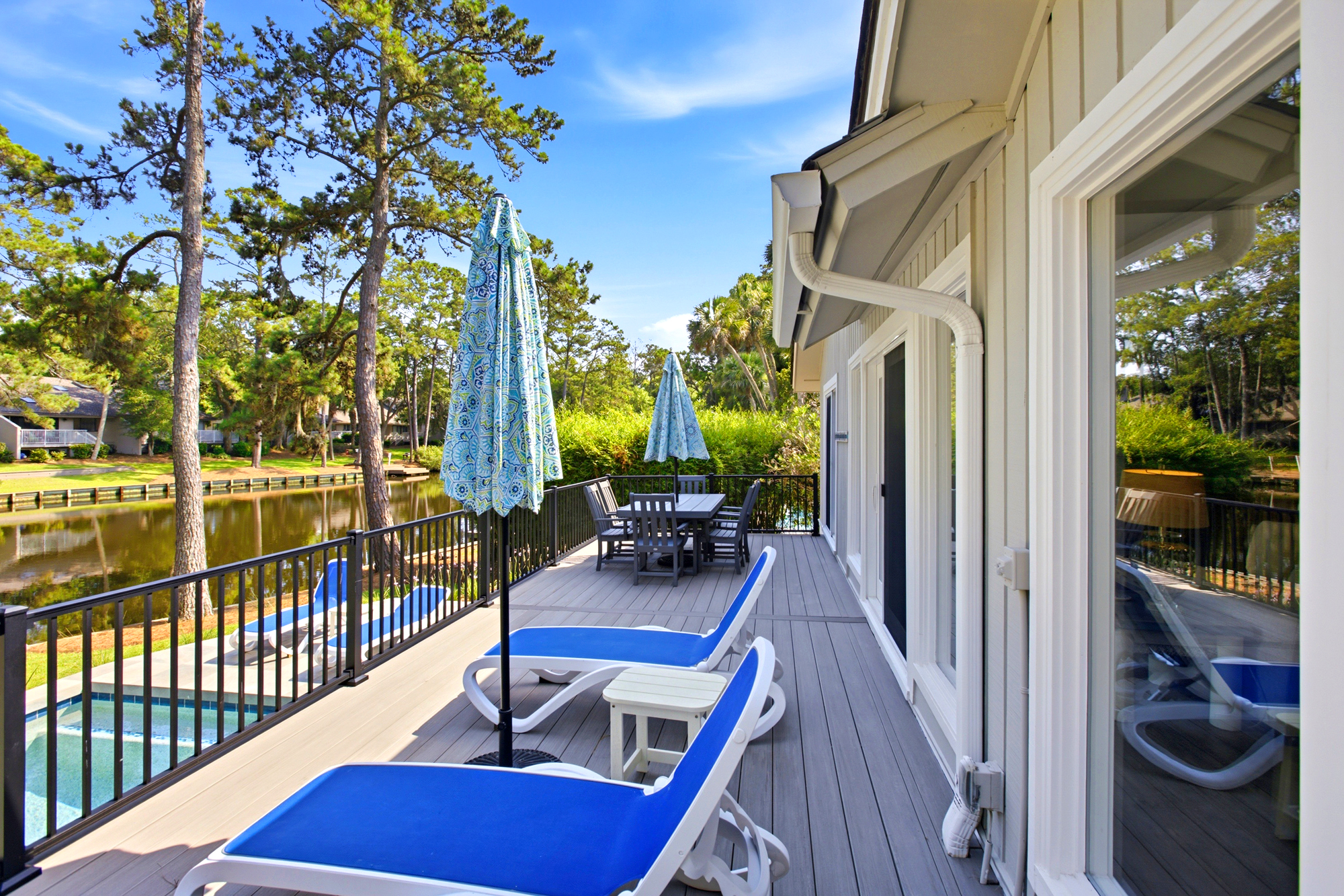 Cobalt loungers line this expansive timber deck, offering a front-row seat to the shimmering canal and swaying pines. Afternoon glow warms the outdoor dining area, creating a serene spot for alfresco meals by the water.
