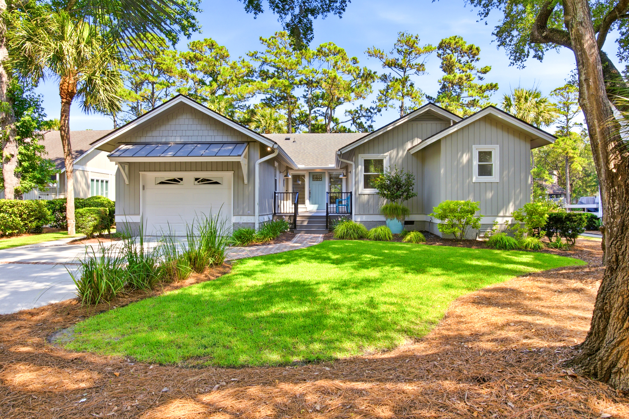 Coastal pines and swaying palms frame this serene cottage, where soft gray siding meets a bright turquoise door. Sun-drenched lawns and a neat brick path create a cheering, quiet frontage for your seaside escape.
