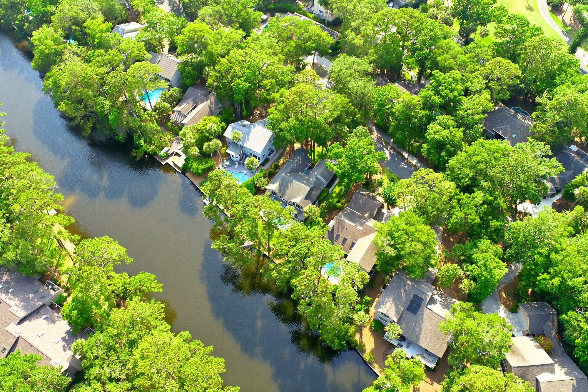 Leafy canopies shade this peaceful waterfront neighborhood, where shingle roofs and private docks meet the calm, dark water. Morning brightness plays across the lush gardens and turquoise pools, creating a quiet sanctuary for your next getaway.