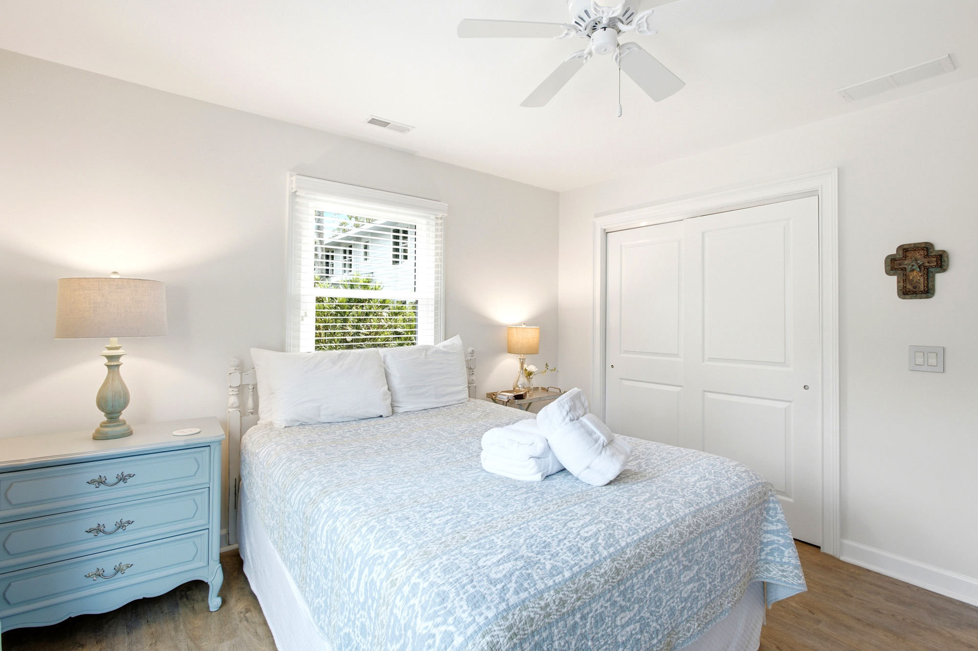 Sky-blue accents and a patterned coverlet create a serene escape, while the morning glow filters through the blinds to illuminate the plush linens. This airy bedroom pairs vintage-style furniture with a bright, clean atmosphere for a peaceful nap.