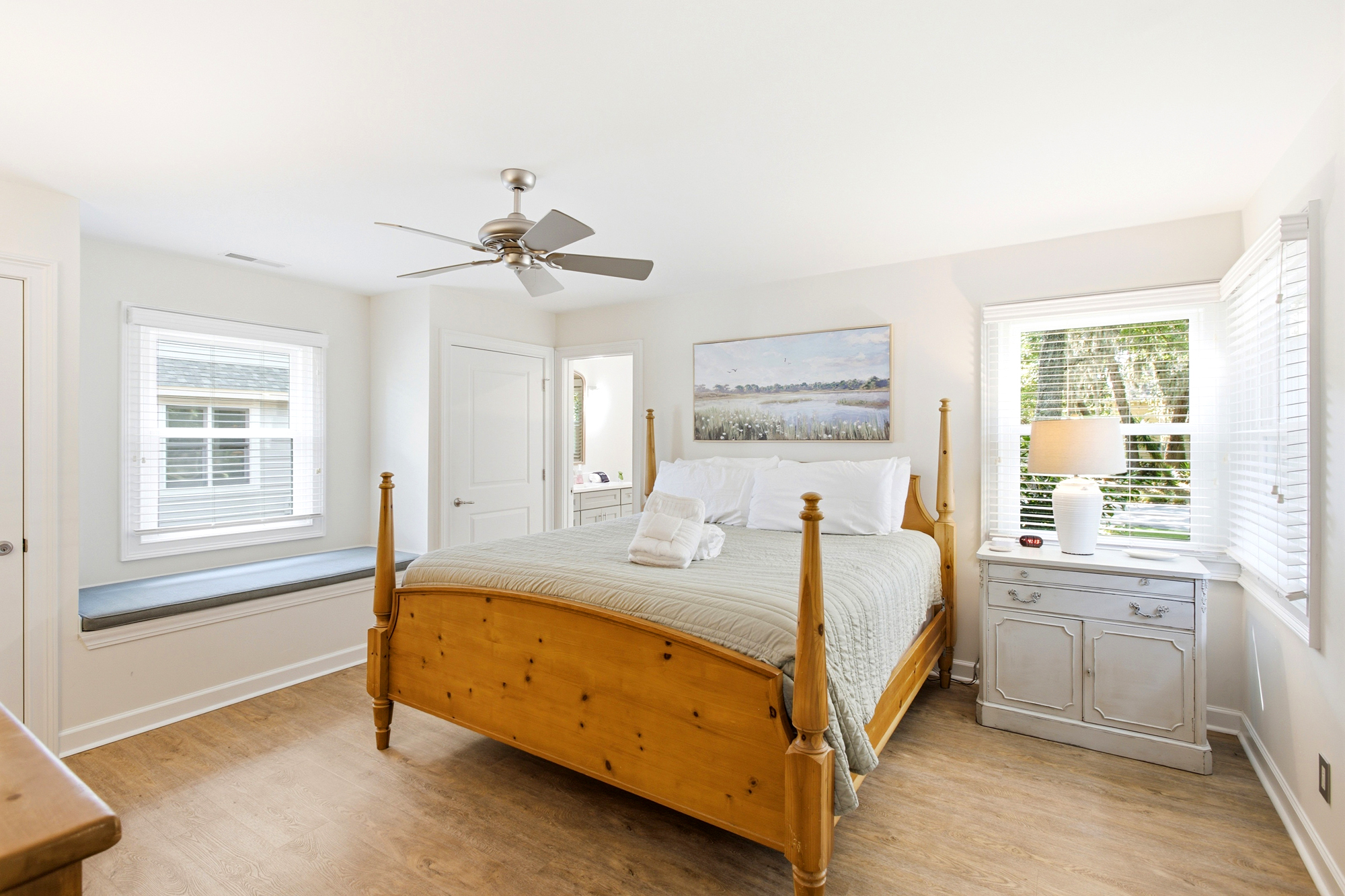 Pine wood grains and peaceful morning light fill this airy bedroom, where a plush window seat invites quiet contemplation. Serene landscape art and cool, quilted textures create a grounding space for a restful night.
