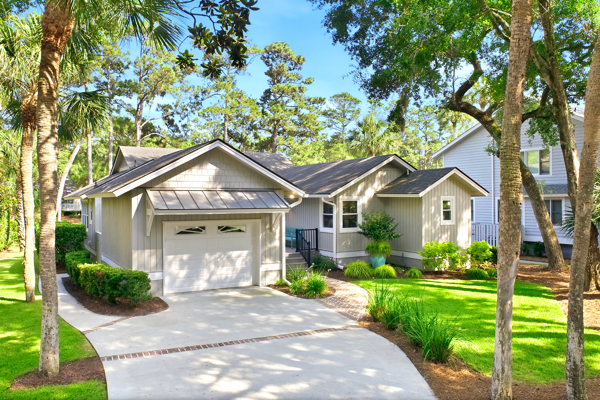 Tall palms and leafy oaks frame this coastal cottage, where soft gray siding meets a lush, sun-drenched lawn. A brick-lined path leads you home through shadows cast by the afternoon glow, promising a quiet and private escape.