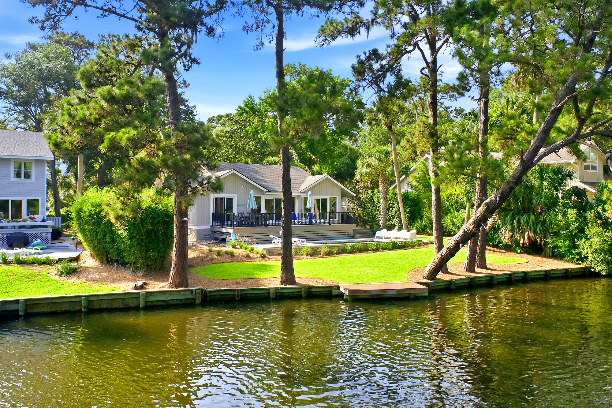 Waterfront living defines this serene scape, where a private dock meets a lush lawn and a cool pool. Slender pines frame the view of the deck, offering a perfect spot to watch the ripples of the canal under the bright afternoon glow.