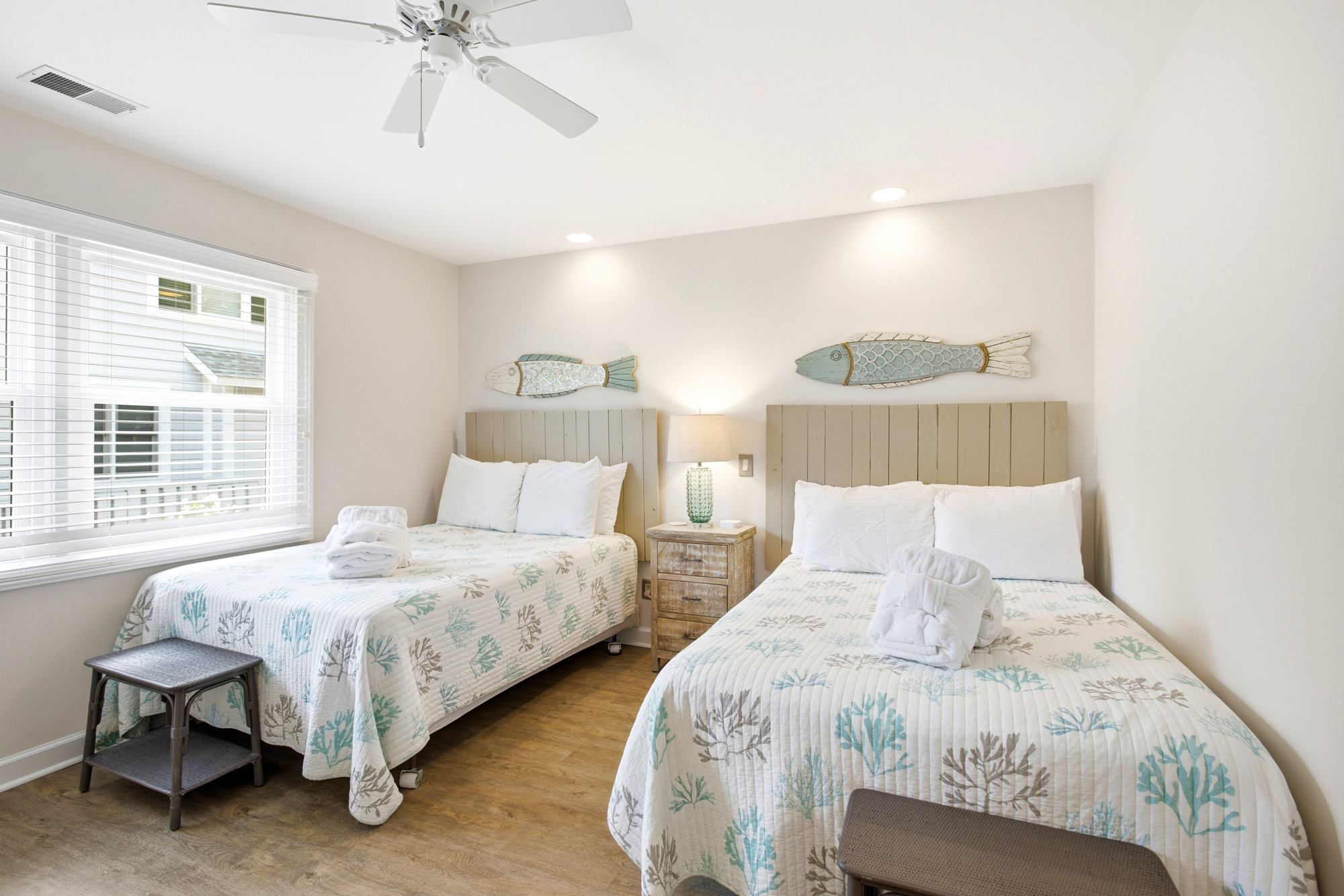 Weathered wood headboards and playful aquatic accents create a serene beachside mood in this airy twin bedroom. Bright day glow pours through the blinds, highlighting the cool textures and fresh linens for a restful night.