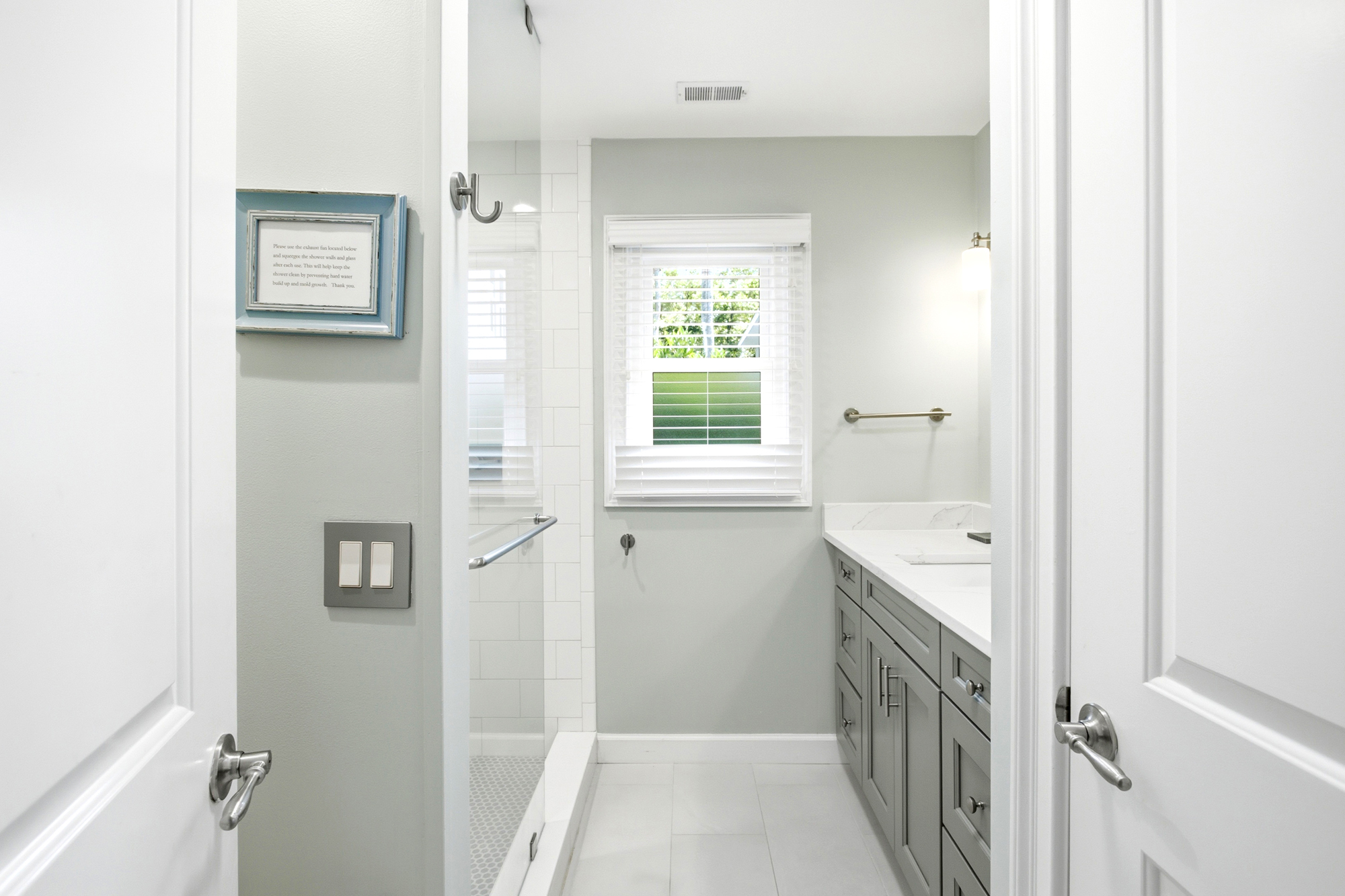 Pale gray cabinetry and smooth marble surfaces create a serene backdrop for your morning routine. Daylight filters through the window, reflecting off the glass shower and clean subway tiles to keep the room feeling airy and bright.