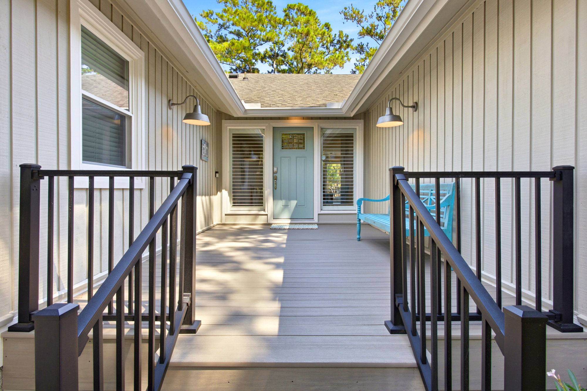 Dappled shadows dance across the wide wooden planks of this serene entryway, where a soft blue door and matching bench offer a peaceful greeting. Industrial sconces glow against neutral siding, framing a path that feels both modern and homey.