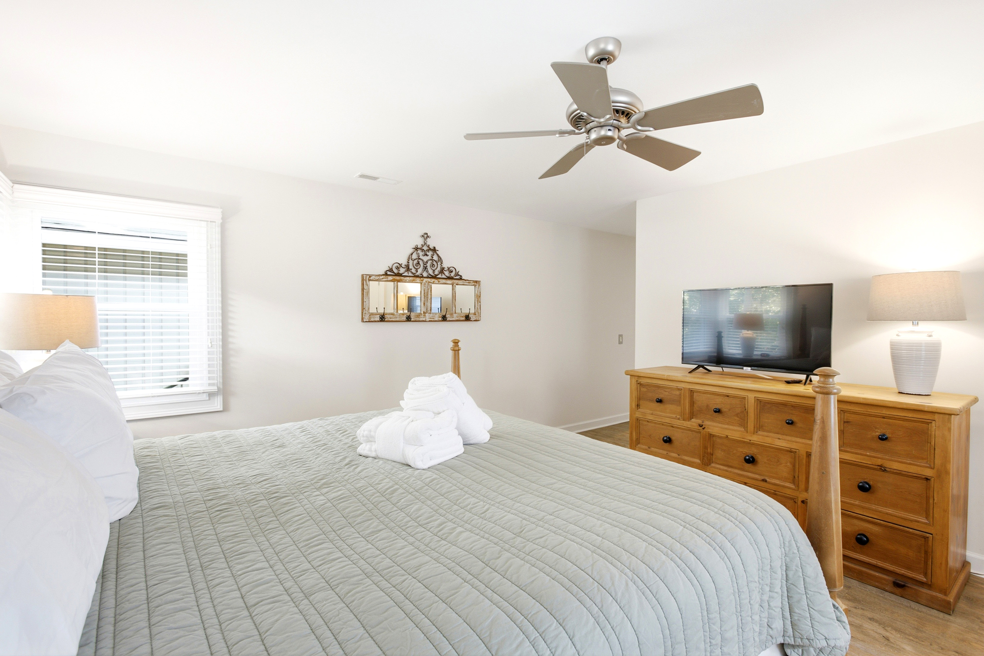 Quilted linens in a calming sage green pair with warm timber furniture to create a serene sanctuary. Morning rays filter through the blinds, catching the metallic sheen of the ceiling fan and the glow of the accent lamps.