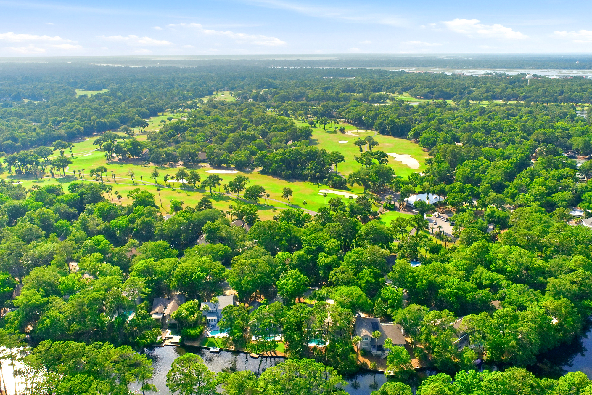 Verdant fairways wind through a dense canopy of ancient oaks, creating a scenic tapestry of green and blue. Sunlit waterways and quiet neighborhood paths invite slow afternoons spent exploring this lush, peaceful landscape from above.