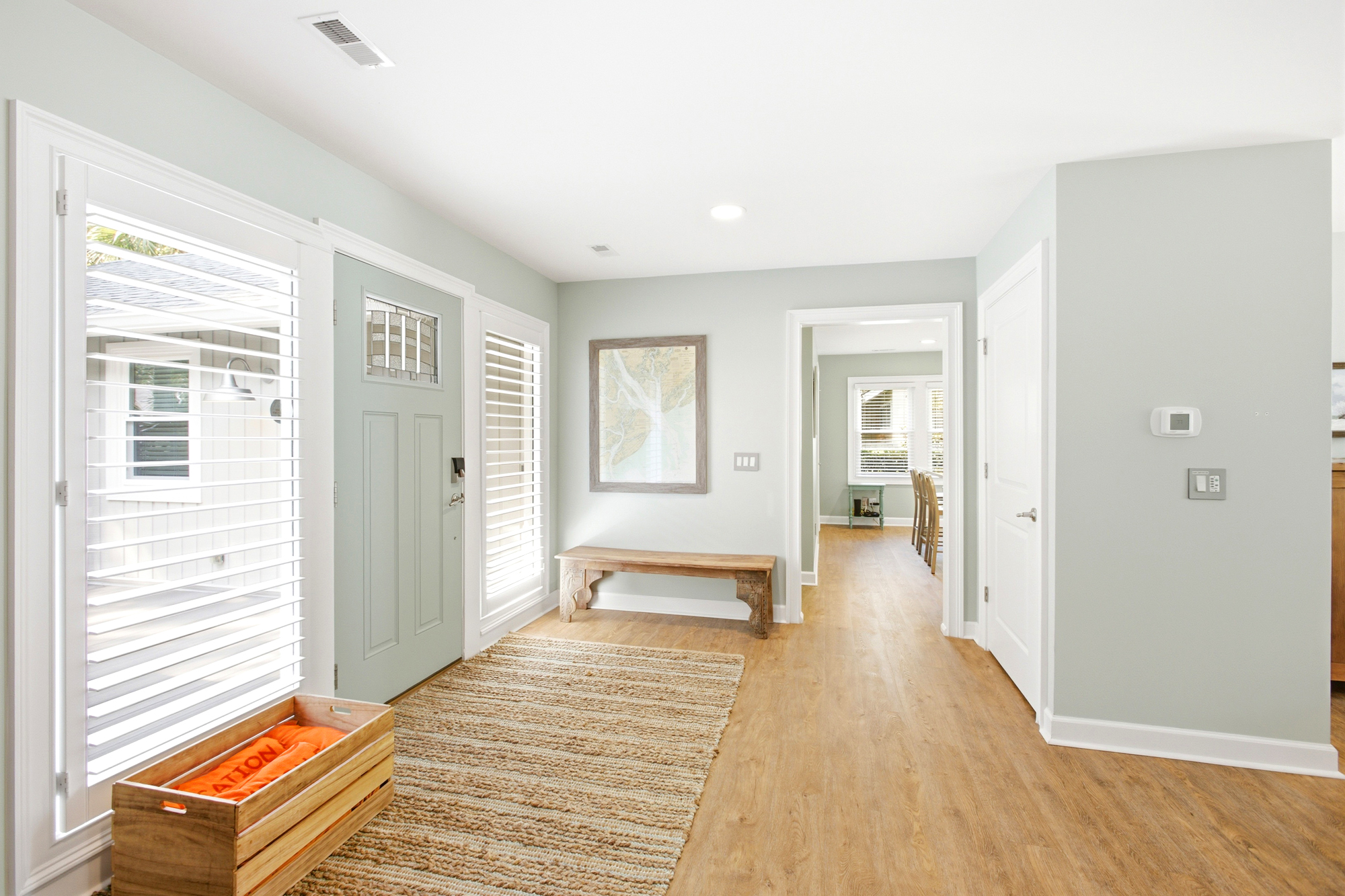 Sage green walls and honey colored floors create a serene entryway filled with crisp morning light. A textured jute rug and rustic wooden bench offer a grounded, organic feel as you move into the airy living space.