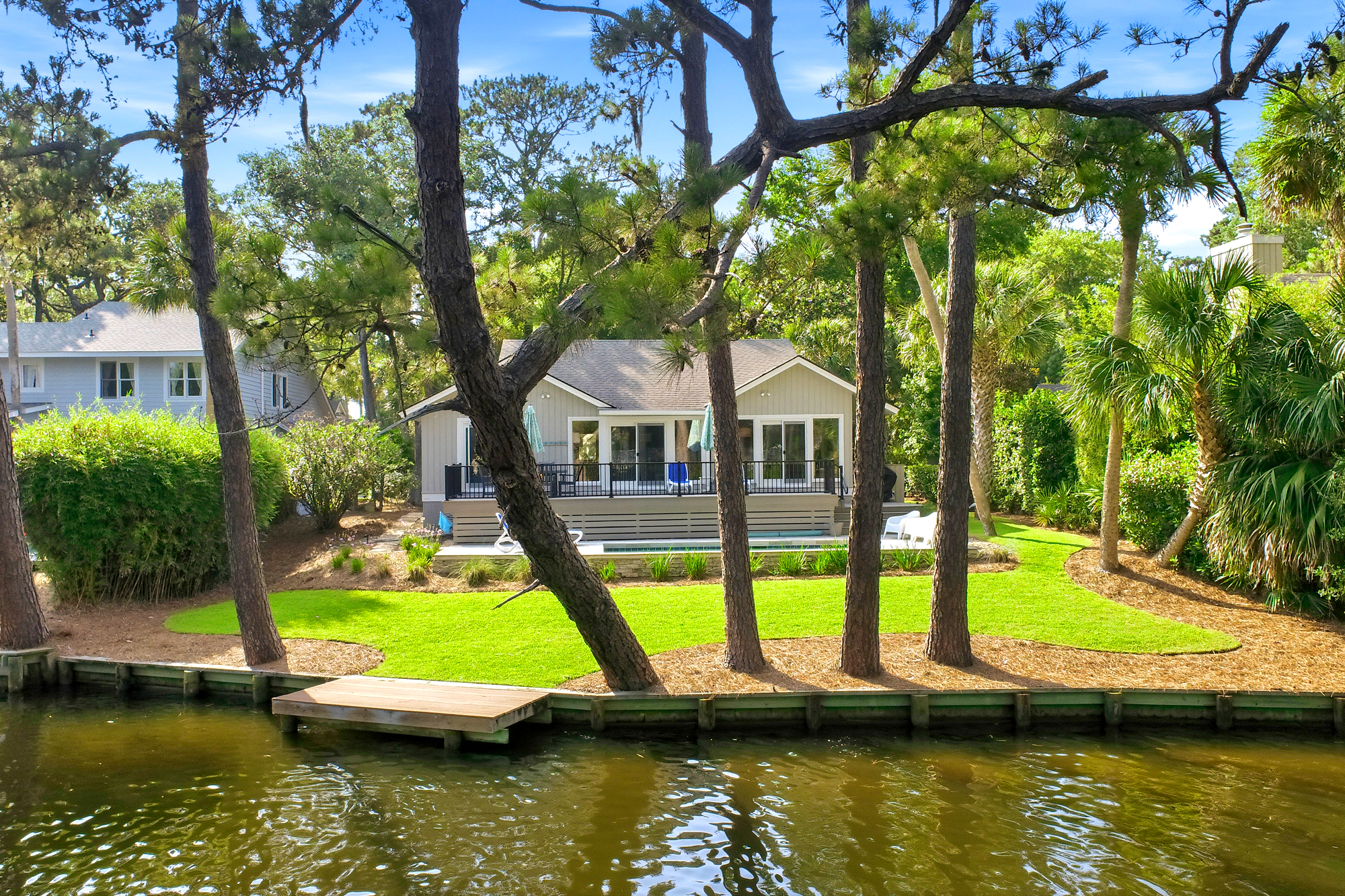 Deep green lawns and sturdy pines frame this quiet canal-side home, where a private wooden dock meets the gentle ripples of the water. Large windows and an expansive deck invite the bright afternoon sky into every corner of the yard.
