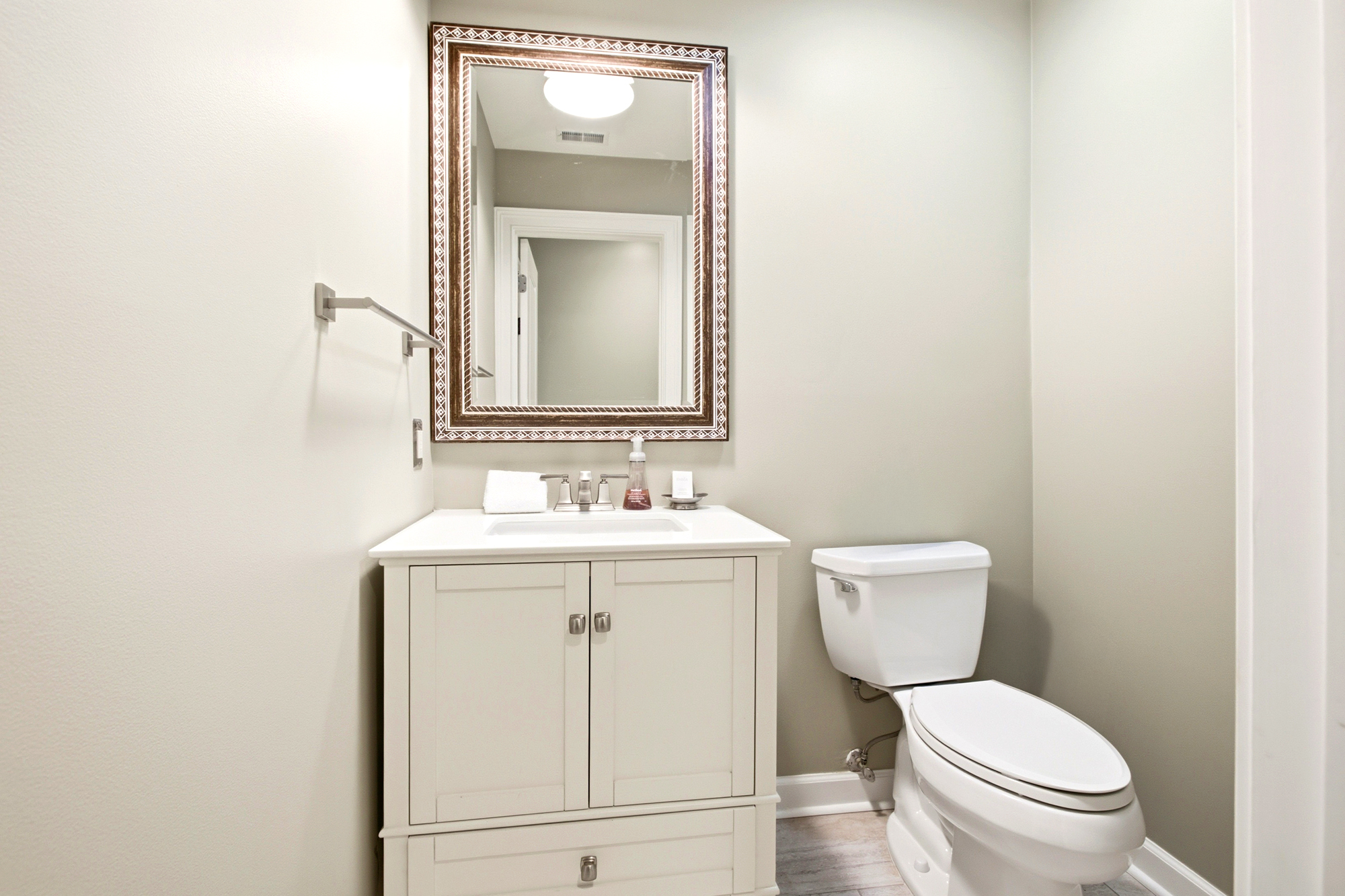 Ornate bronze frames a large mirror, adding a touch of classic elegance to this bright and clean powder room. Soft cream cabinetry and muted walls create a serene, quiet atmosphere for your morning routine.