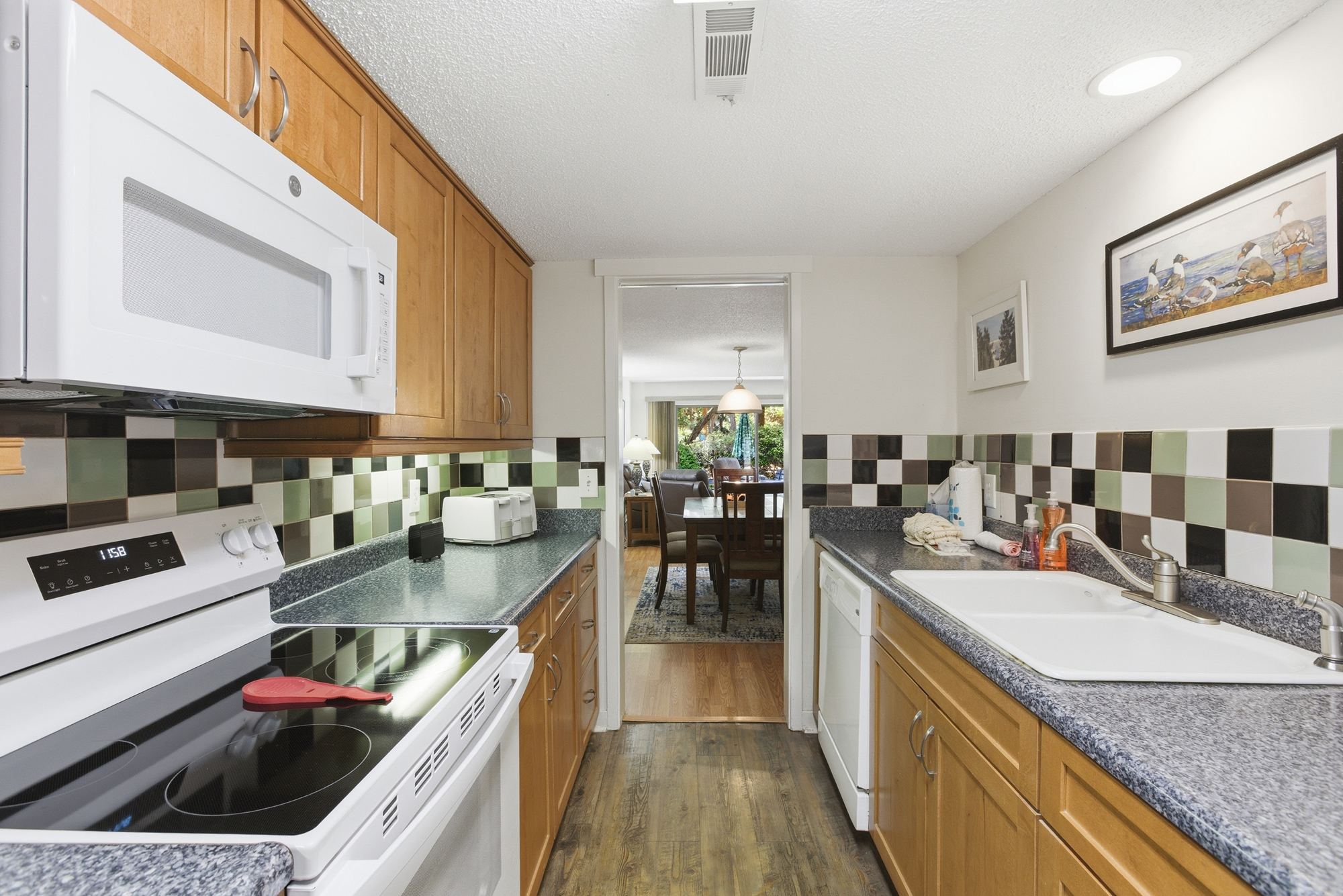 Chequered tiles in earthy tones bring a playful character to this efficient kitchen, where warm wood cabinetry meets speckled stone counters. Bright overhead lighting illuminates the path toward a cozy dining area and a view of the garden beyond.