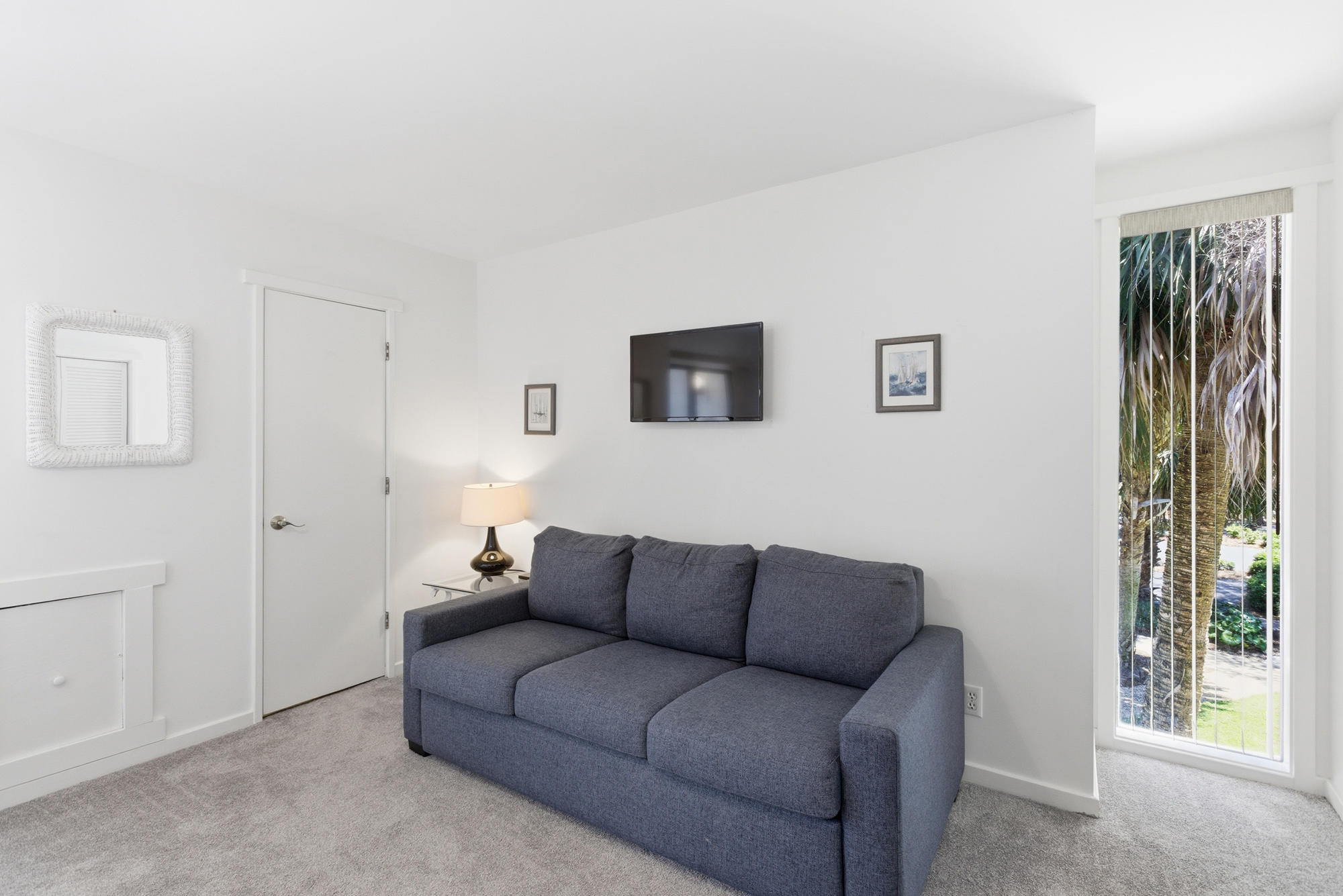Denim blue upholstery anchors this serene lounge, where floor to ceiling glass invites views of tropical fronds inside. Soft carpet and a glowing lamp create a quiet corner for movie nights in this airy, bright space.